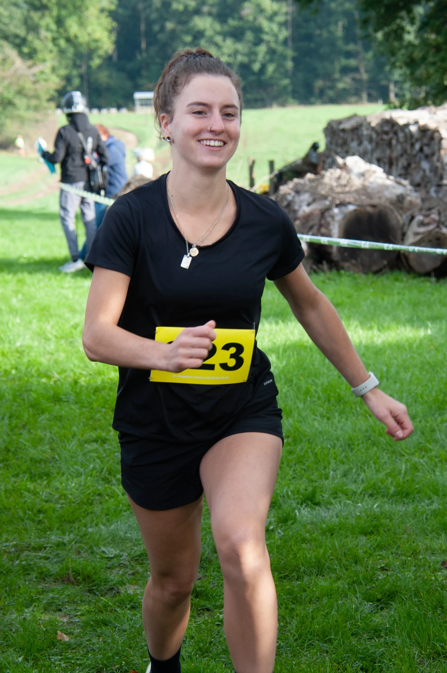 Une femme souriante en train de courir lors d'une course en plein air, portant un t-shirt noir, un short noir, une montre et une plaque de course avec le numéro 23.
