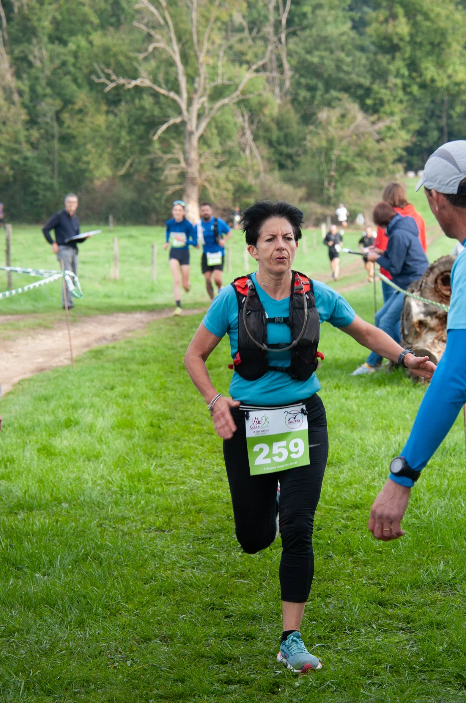 Une femme court dans un événement sportif en plein air, portant un dossard numéro 259, une bouteille d'eau autour du cou, et une veste de course. Elle semble concentrée, avec un groupe de personnes en arrière-plan dans un environnement naturel avec d