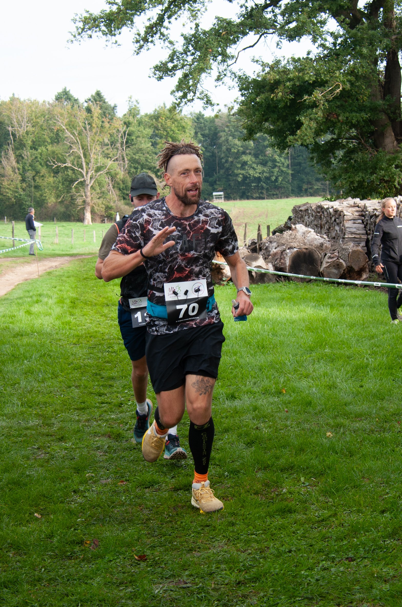 Un homme court lors d'une compétition en plein air, portant un maillot à motif foncé, un numéro 70, et des vêtements de course, avec une surface herbeuse et des arbres verts en arrière-plan.