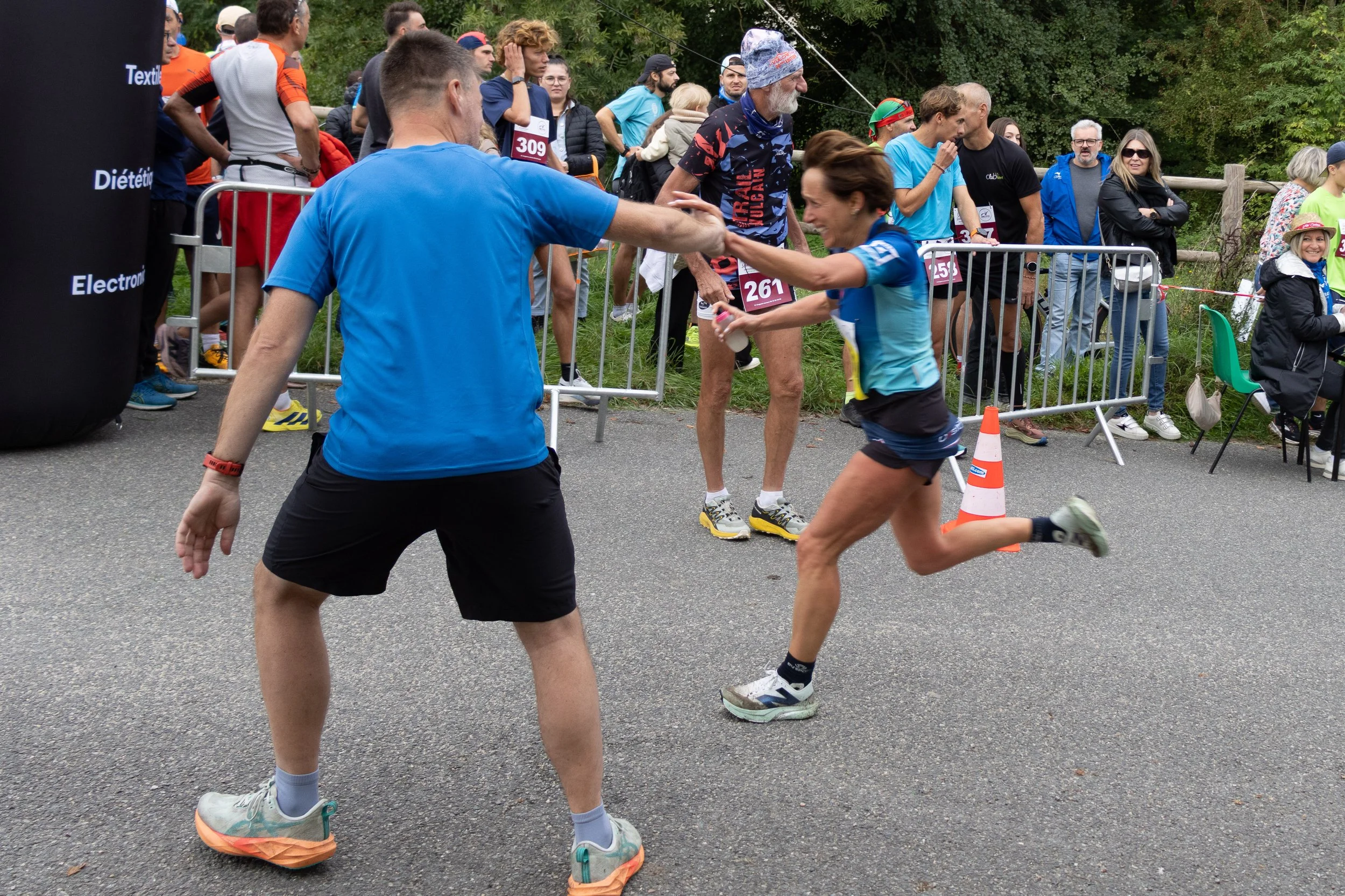Un homme en bleu tend la main à une femme en course lors d'une compétition de course à pied, avec un groupe de spectateurs derrière une barrière.