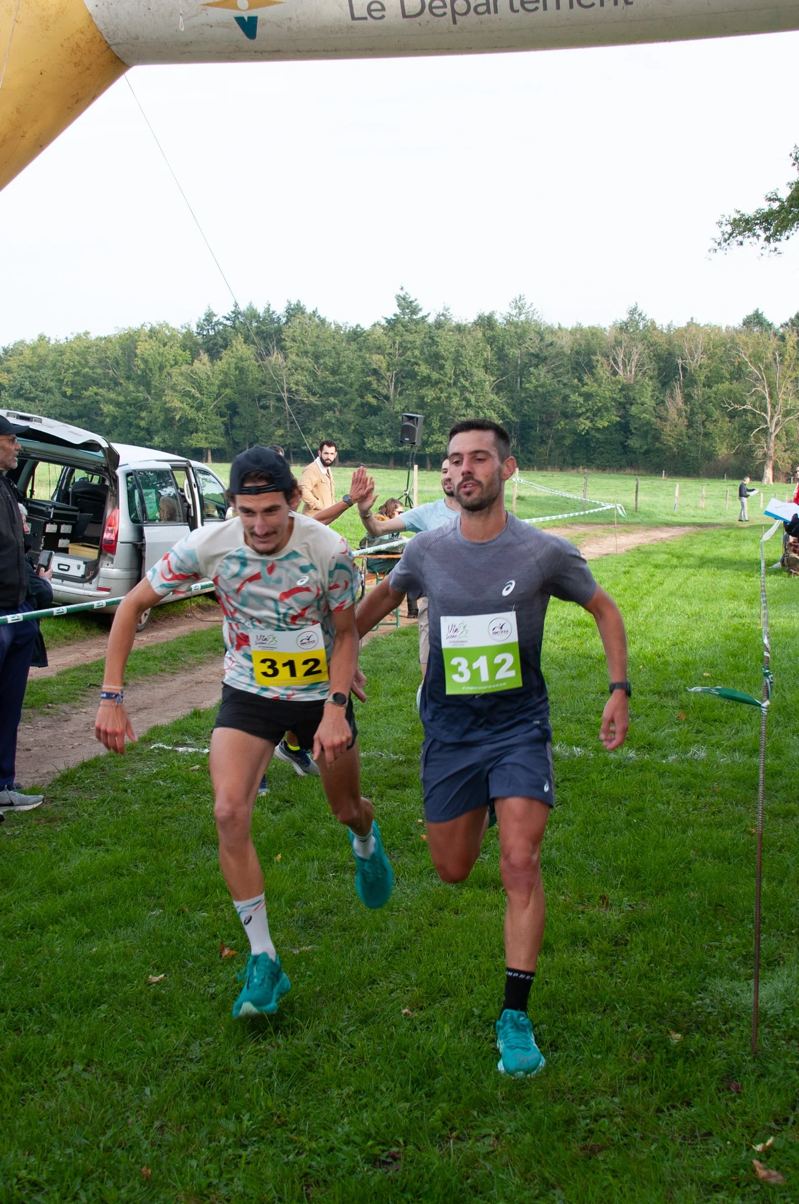 Deux coureurs franchissent la ligne d'arrivée lors d'une course en plein air, portant des dossards numérotés 312, avec un environnement naturel en arrière-plan et des personnes en arrière-plan célébrant.