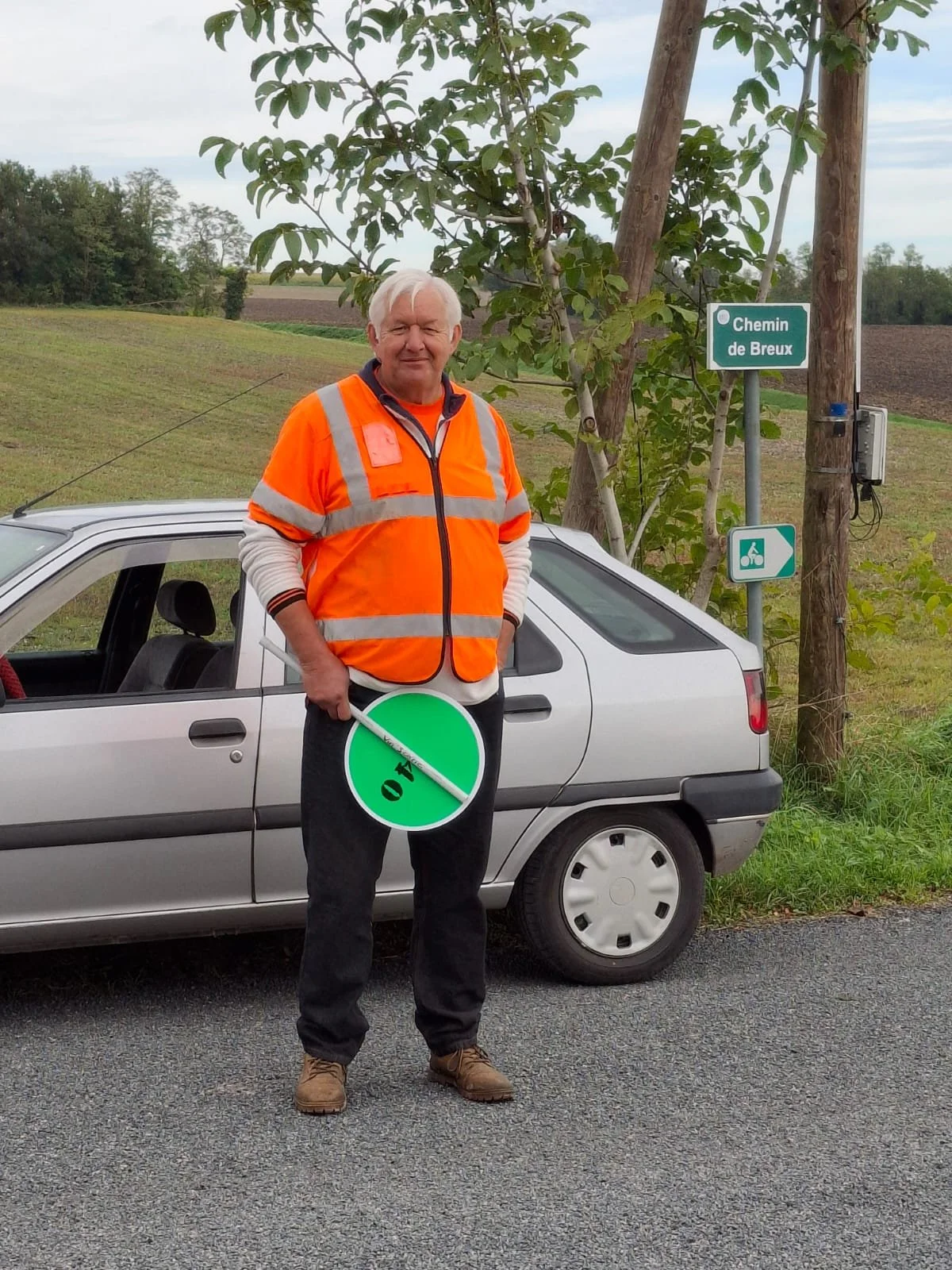 Un homme en veste orange de sécurité tient un panneau de limite de vitesse de 40 km/h. Il se tient devant une voiture grise sur le bord d'une route rurale avec des champs et des arbres en arrière-plan. Il y a un panneau indiquant 'Chemin de Breux' et
