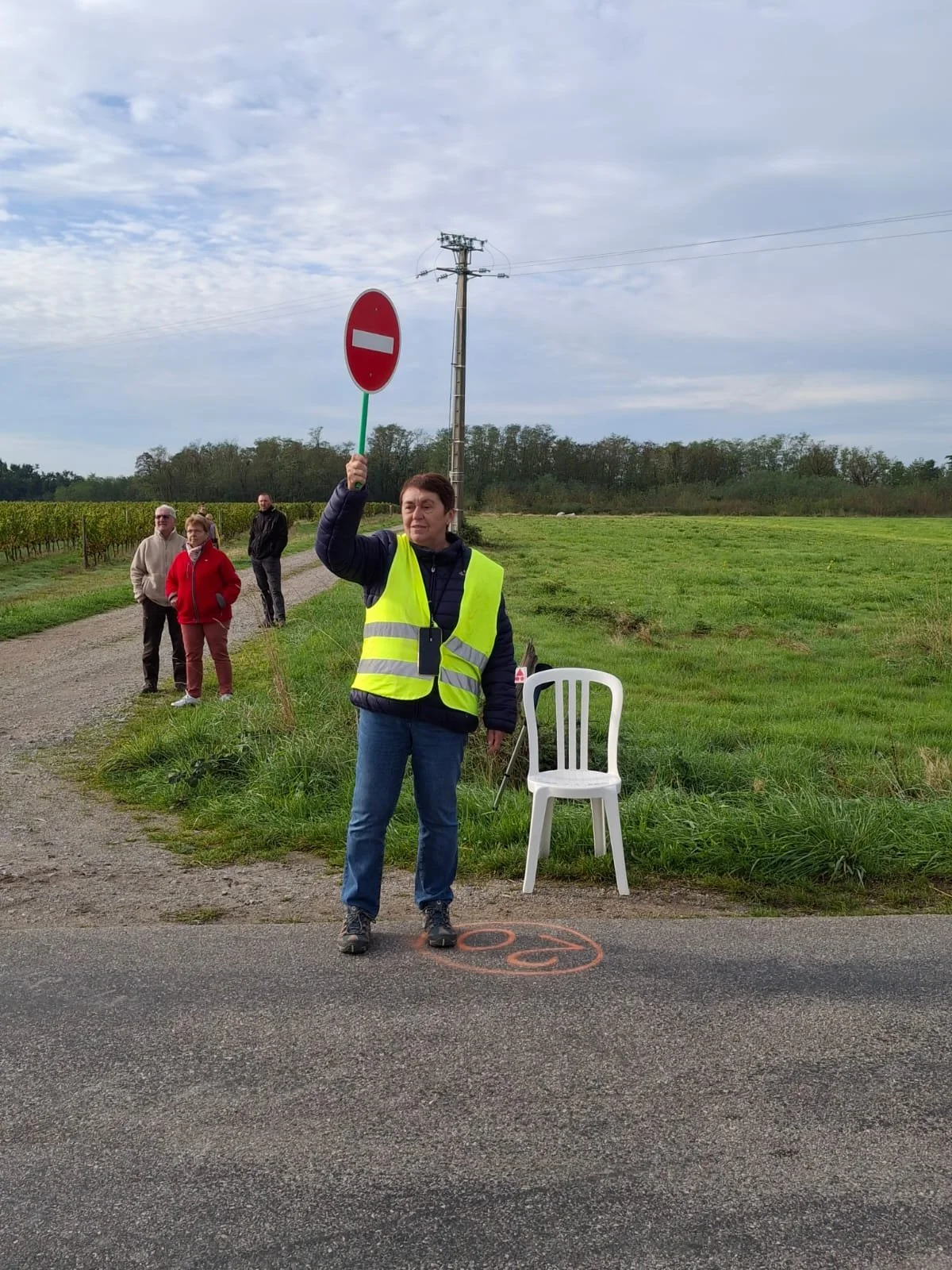 Un homme portant un gilet jaune de sécurité tient un panneau d'interdiction sur une route rurale, avec d'autres personnes en arrière-plan dans un paysage agricole.