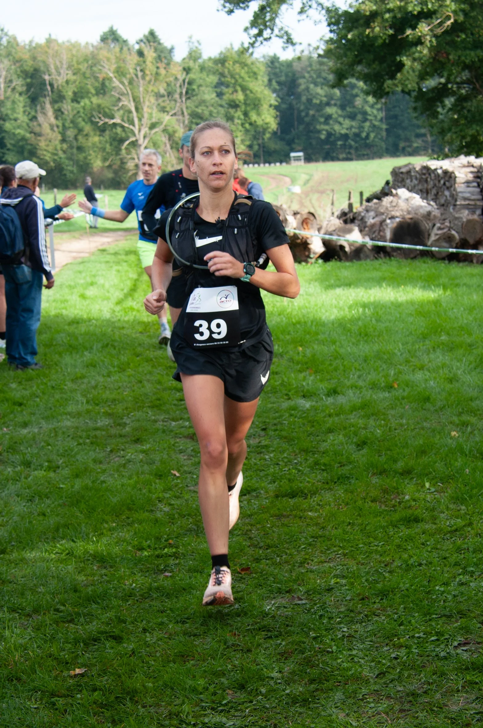 Une femme court lors d'une course en plein air, portant un maillot noir avec le numéro 39, entourée de spectateurs et d'une nature verdoyante.