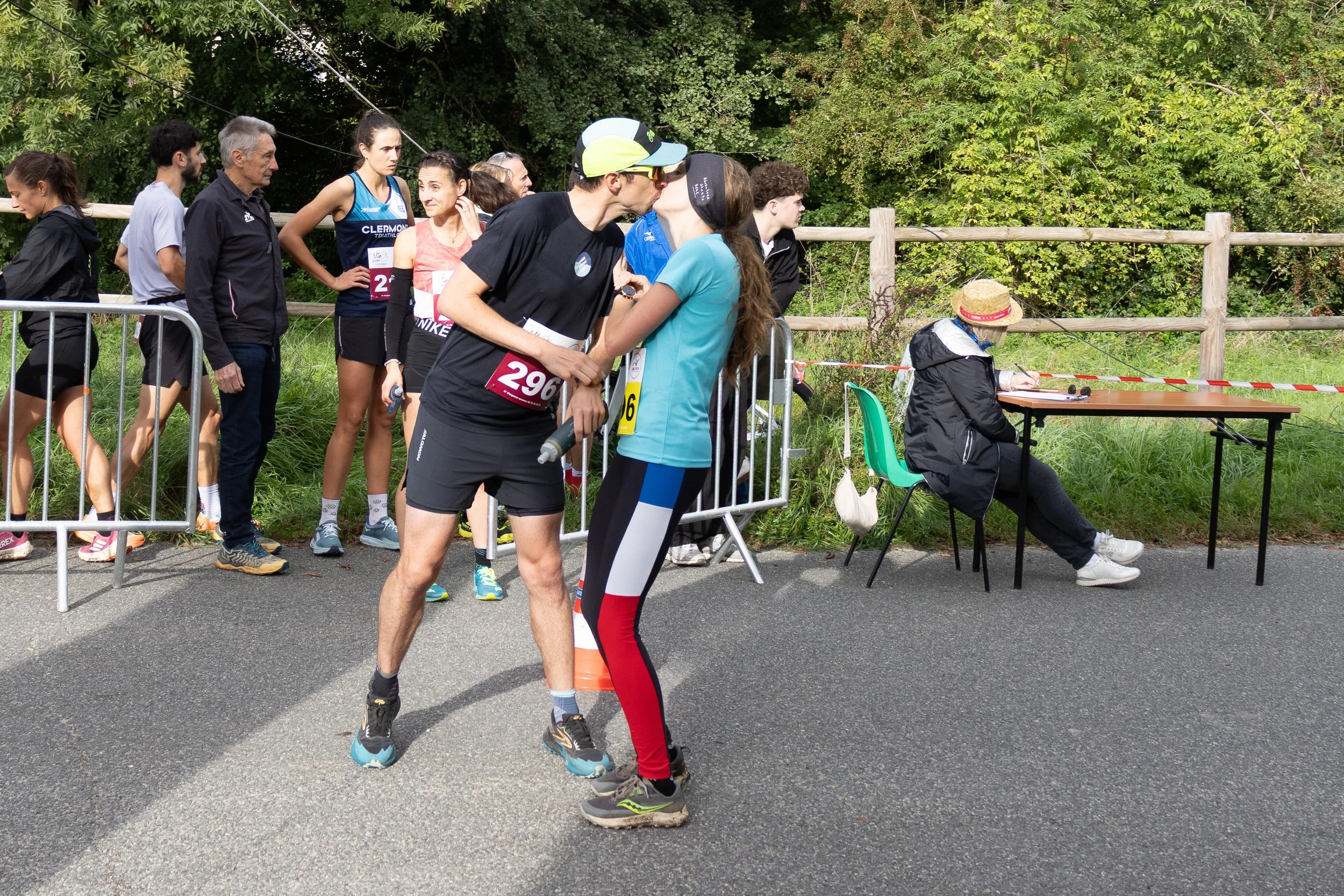 Deux coureurs de marathon s'embrassent devant la ligne d'arrivée, entourés d'un groupe de spectateurs et d'une femme assise à une table près d'une barrière de COVID, avec des arbres en arrière-plan.