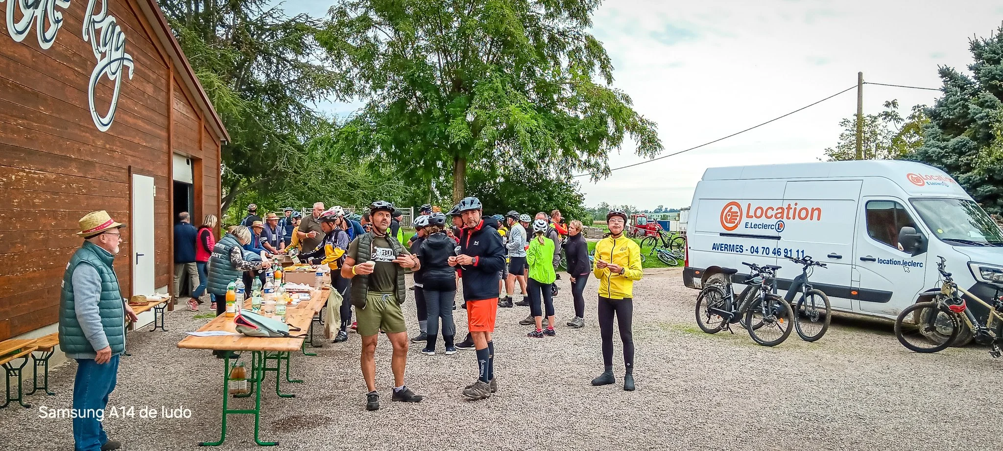 Groupe de cyclistes et de personnes rassemblées devant une offre de boissons et de nourriture à l'extérieur d'un bâtiment en bois, avec des vélos stationnés à côté d'une camionnette blanche de location de vélos. Arbres verts et ciel nuageux en arrière-plan.