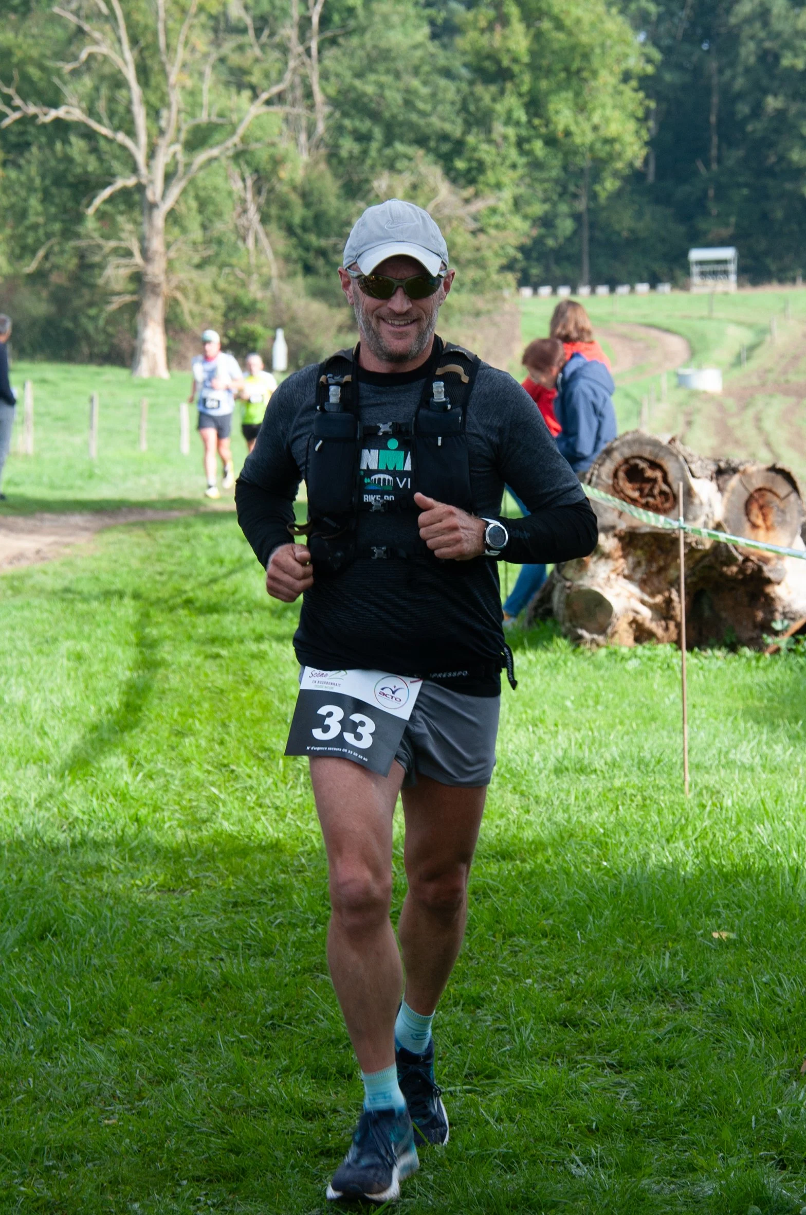 Un homme court lors d'une course en plein air, portant une casquette, des lunettes de soleil, un t-shirt noir, un short gris, et un sac à dos, sur un parcours herbeux entouré d'arbres.
