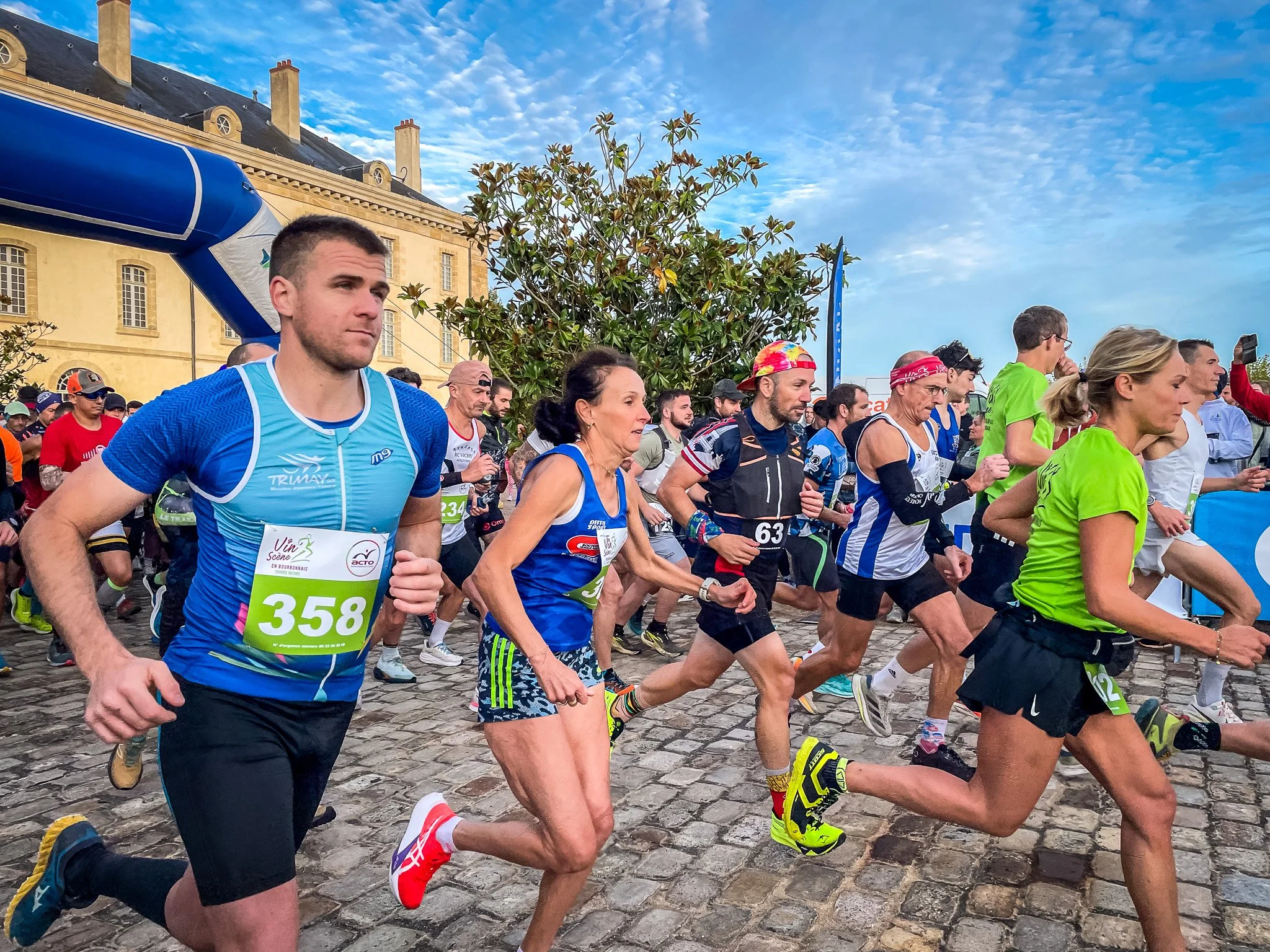 Groupe de coureurs participant à un marathon urbain sur un chemin en pavés, avec un bâtiment ancien en arrière-plan et un ciel bleu