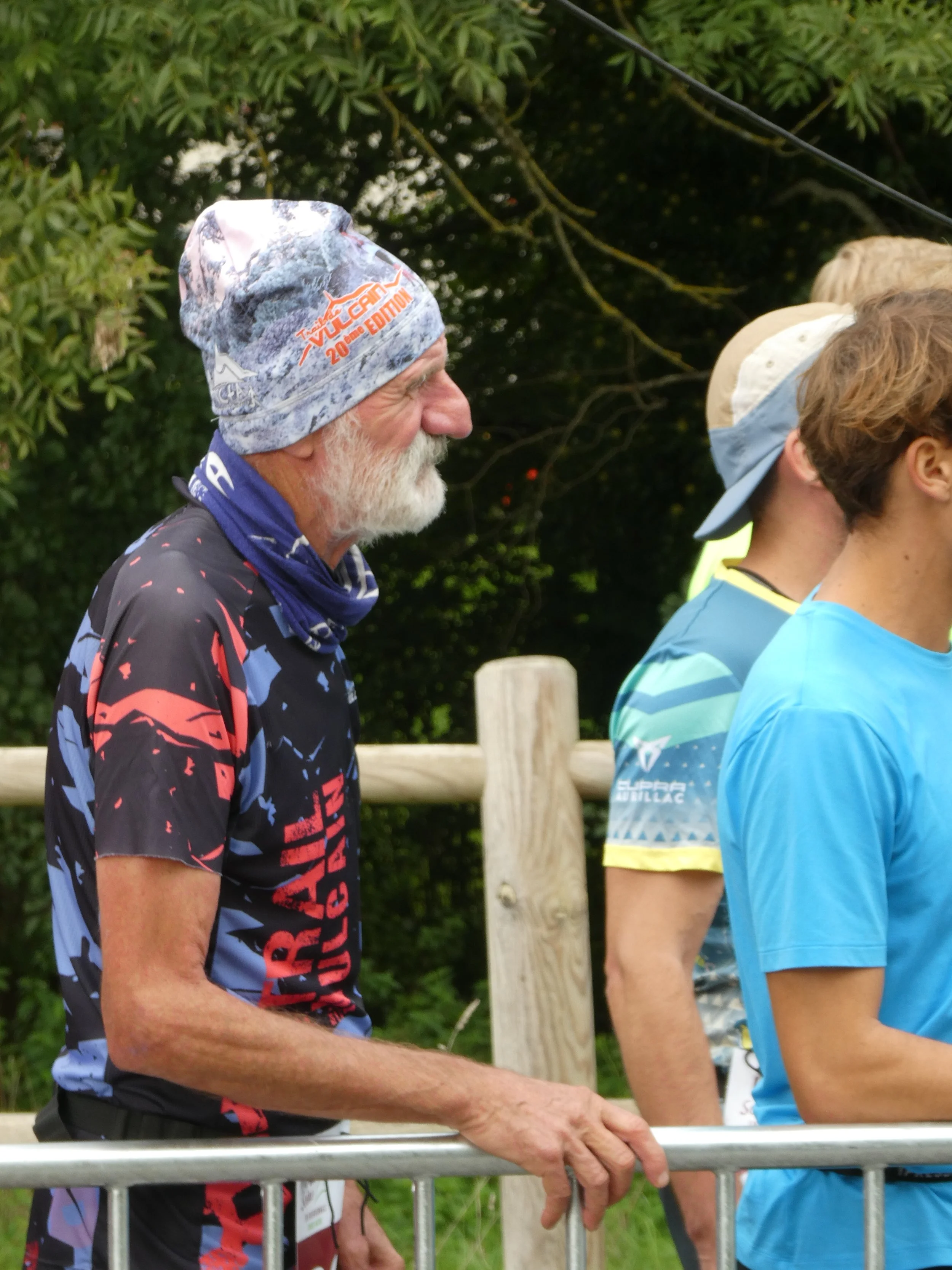 Un homme âgé avec une barbe blanche porte un bonnet de course et un maillot coloré, debout à côté d'une barrière en métal lors d'une course de trail ou de course en plein air.