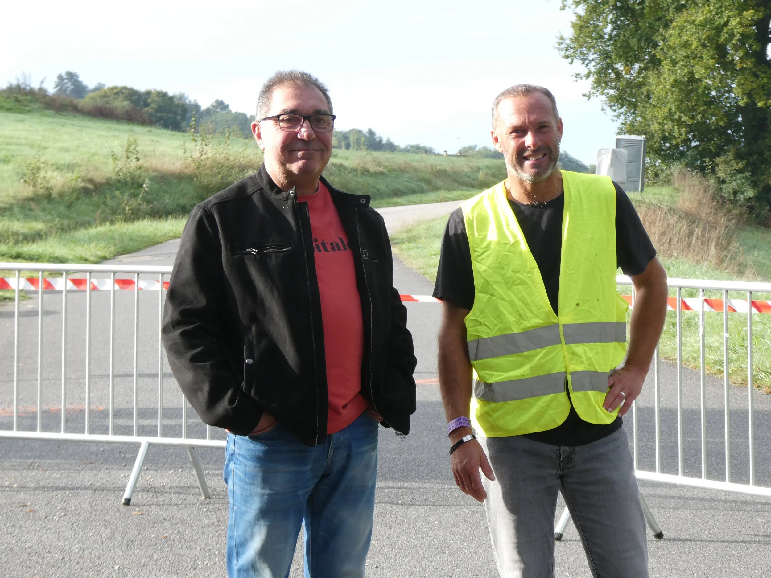 Deux hommes souriants, l'un en veste noire et t-shirt rose, l'autre en gilet de sécurité jaune, se tiennent devant une barrière en métal sur une route de campagne.