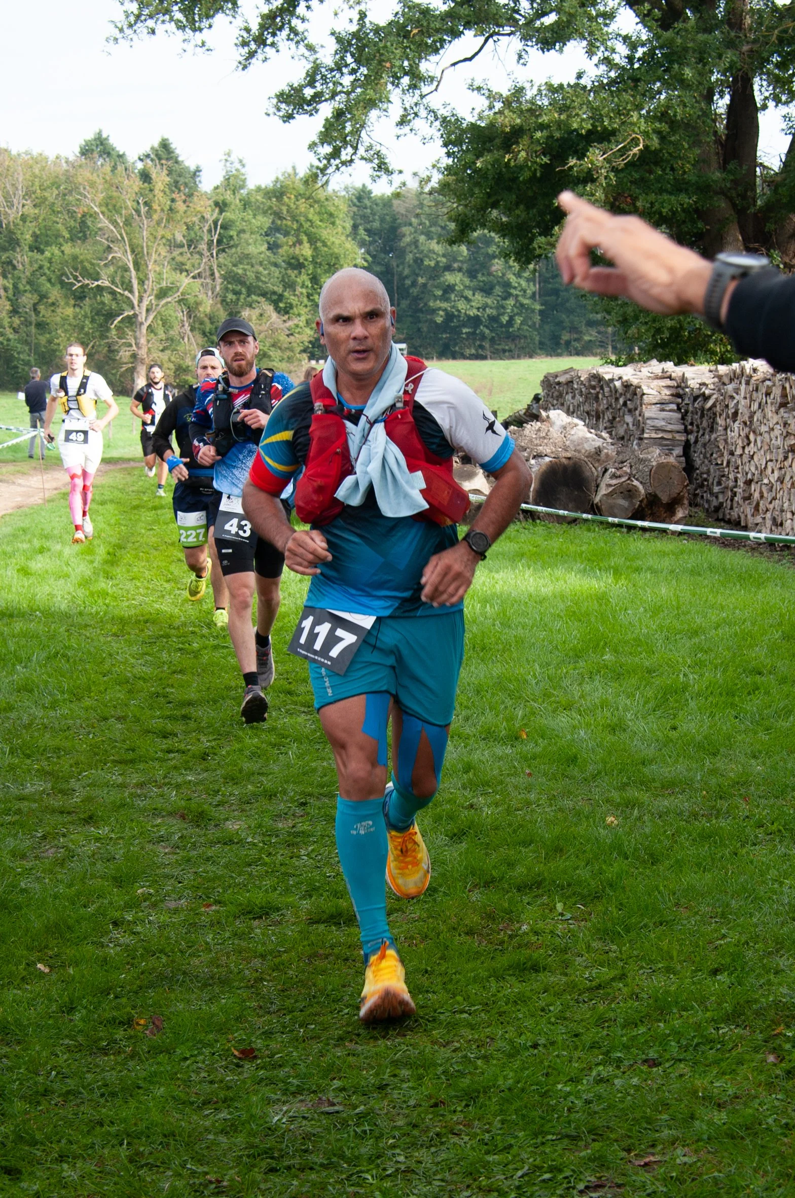 Un groupe de coureurs en compétition dans un parc vert, avec un homme en avant portant un numéro 117, un peu surpris ou concentré.