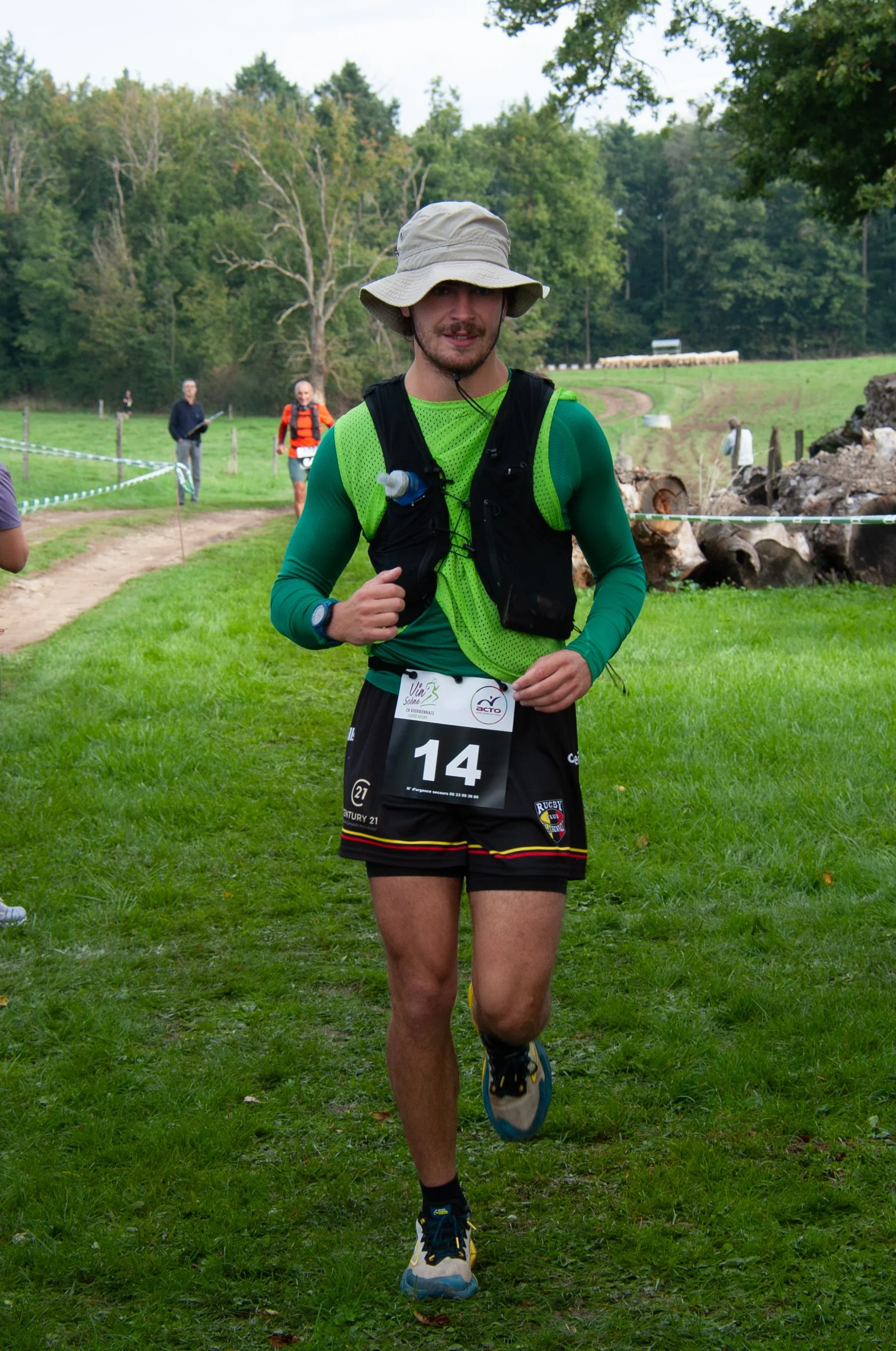 Un homme court dans un trail, portant un chapeau beige, un t-shirt à manches longues vert, une veste de course noire, un dossard numéro 14, et un sac d'hydratation, dans un paysage verdoyant.
