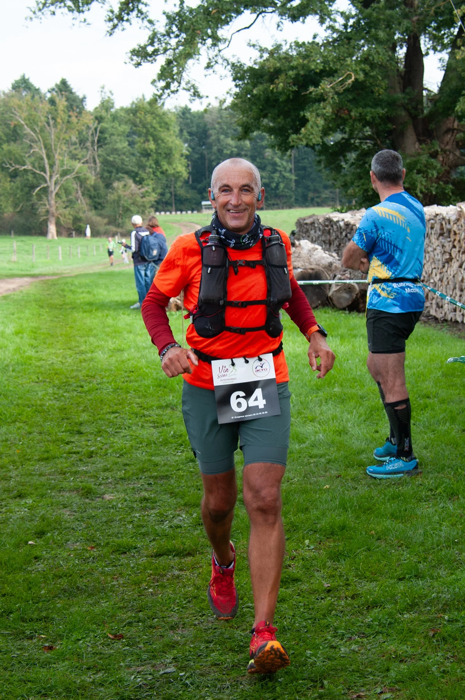Un homme souriant en course lors d'une compétition en plein air, portant un numéro 64, une veste orange, et un équipement de trail. Il y a d'autres coureurs en arrière-plan dans un environnement verdoyant.
