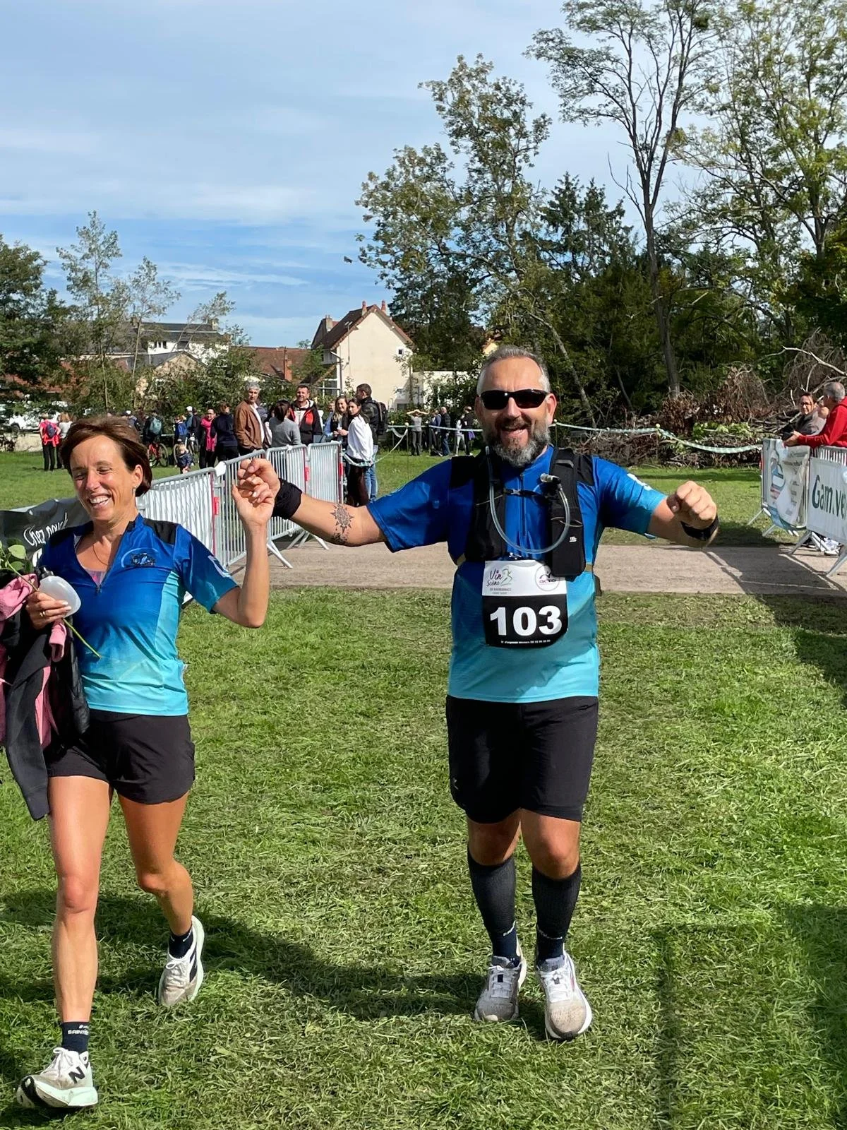 Deux coureurs, un homme et une femme, souriants, en train de finir une course dans un parc avec des spectateurs en arrière-plan. Ils portent tous deux des vêtements de course bleus.