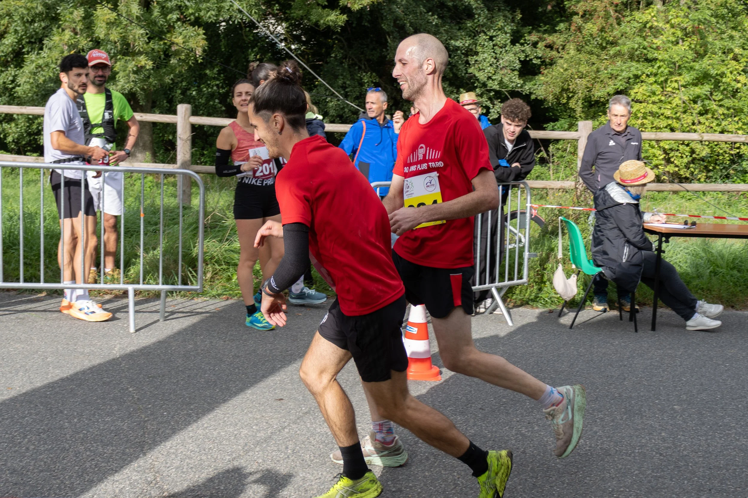 Deux coureurs terminent une course, entourés de spectateurs, certains prennent des photos ou regardent, dans une zone en plein air avec des arbres en arrière-plan.