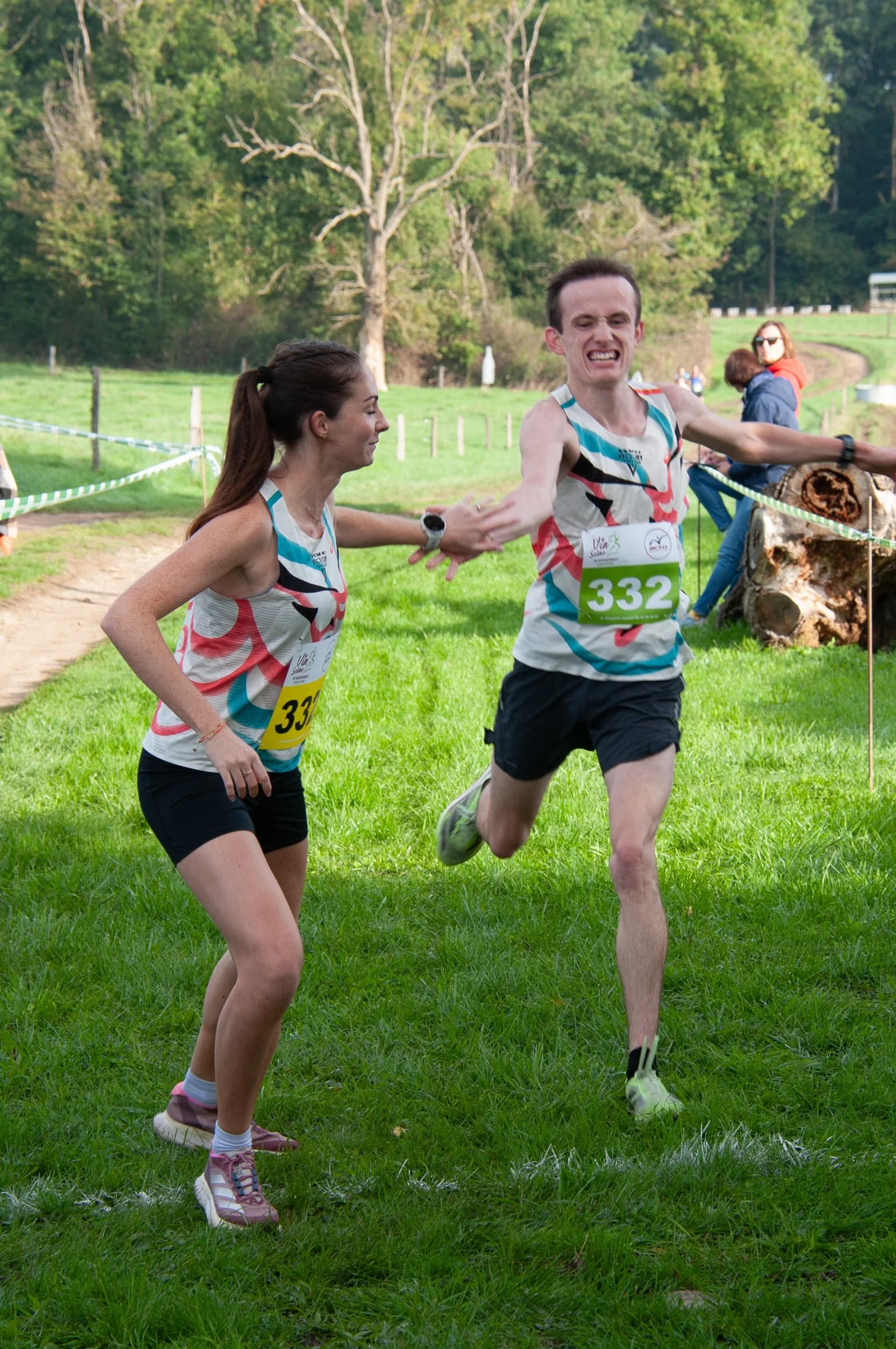 Deux coureurs masculins et féminins se félicitent à la fin d'une course en extérieur. Ils portent des dossards avec les numéros 332 et 33, sur un terrain en herbe avec des arbres en arrière-plan.
