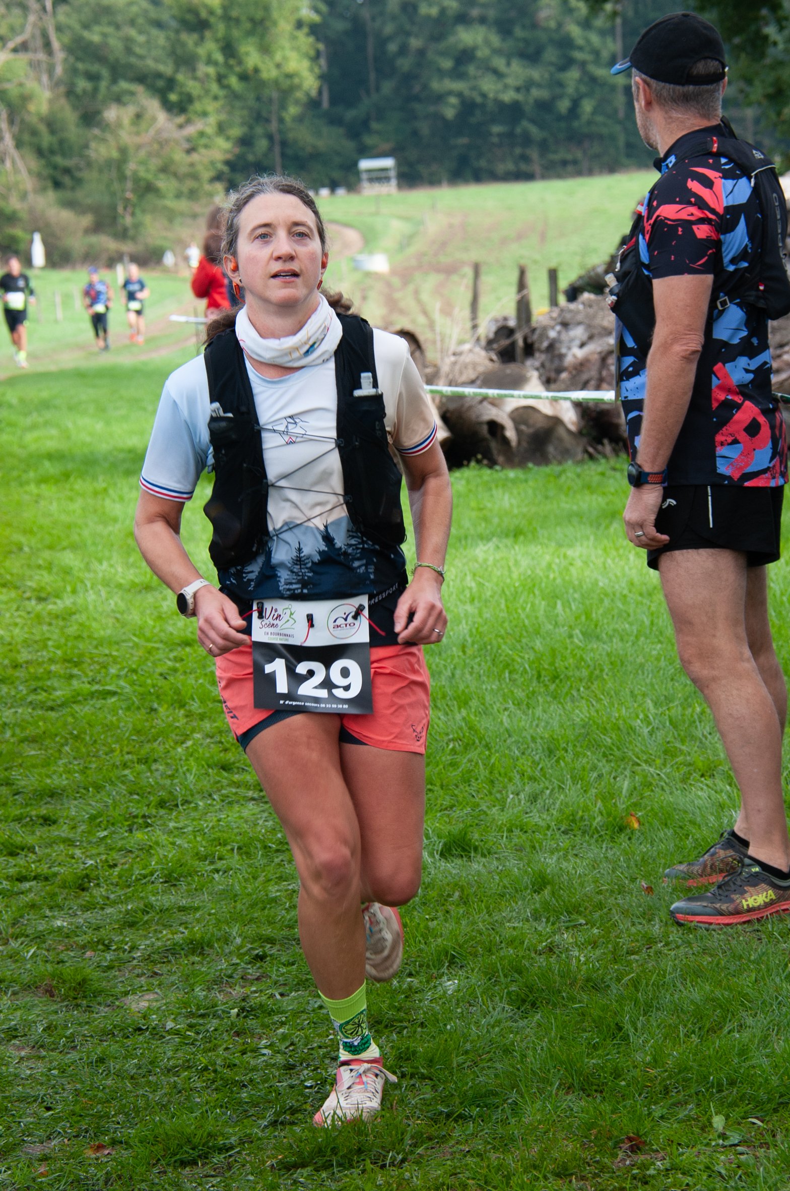 Femme courrant lors d'une course dans un environnement naturel verdoyant, portant un numéro 129, un t-shirt blanc et un short rose, avec un poste d'hydratation à proximité.