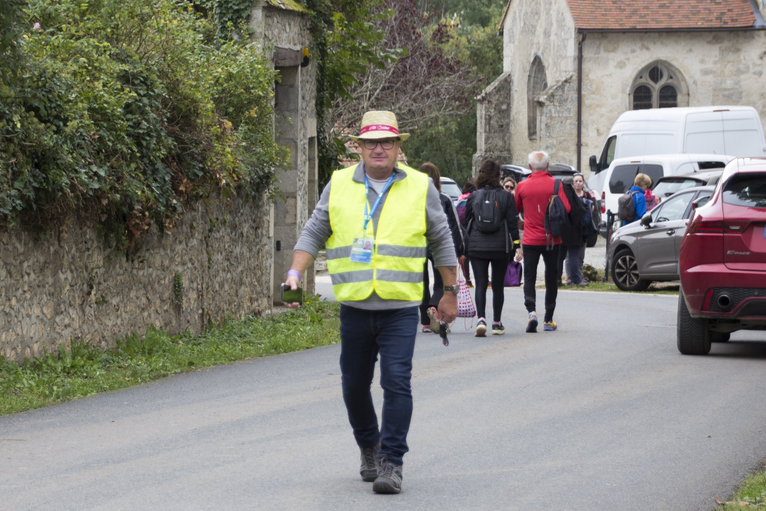 Un homme portant un gilet jaune fluo et un chapeau marche sur une route bordée de maisons en pierre, avec un groupe de personnes en arrière-plan.