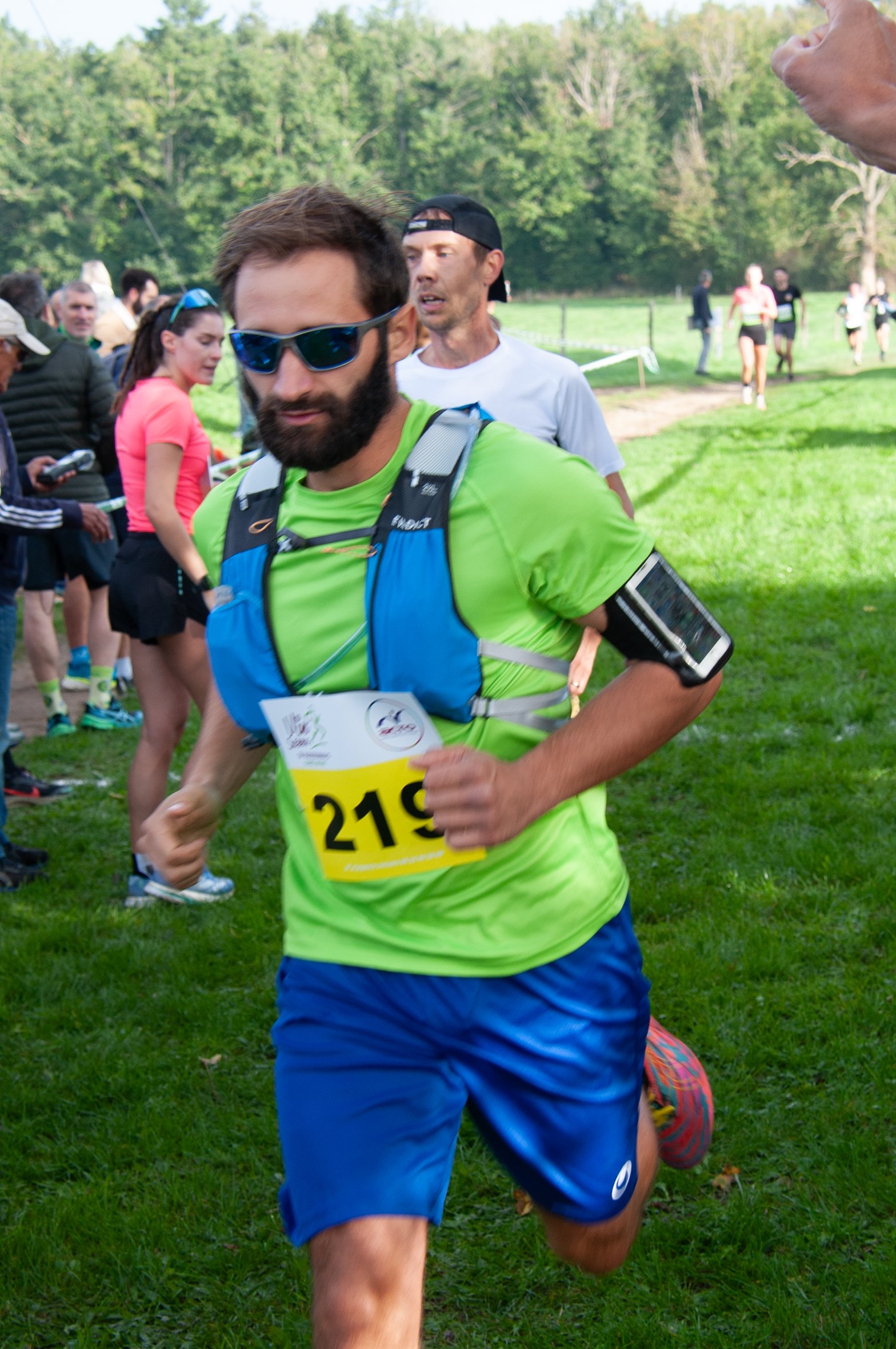 Un homme court lors d'une course en plein air, portant un t-shirt vert, un short bleu, une montre de sport, un bib de course numéro 219, et un bandeau ou bracelet noir. Il porte également un sac à dos et des lunettes de soleil, avec un petit téléphon