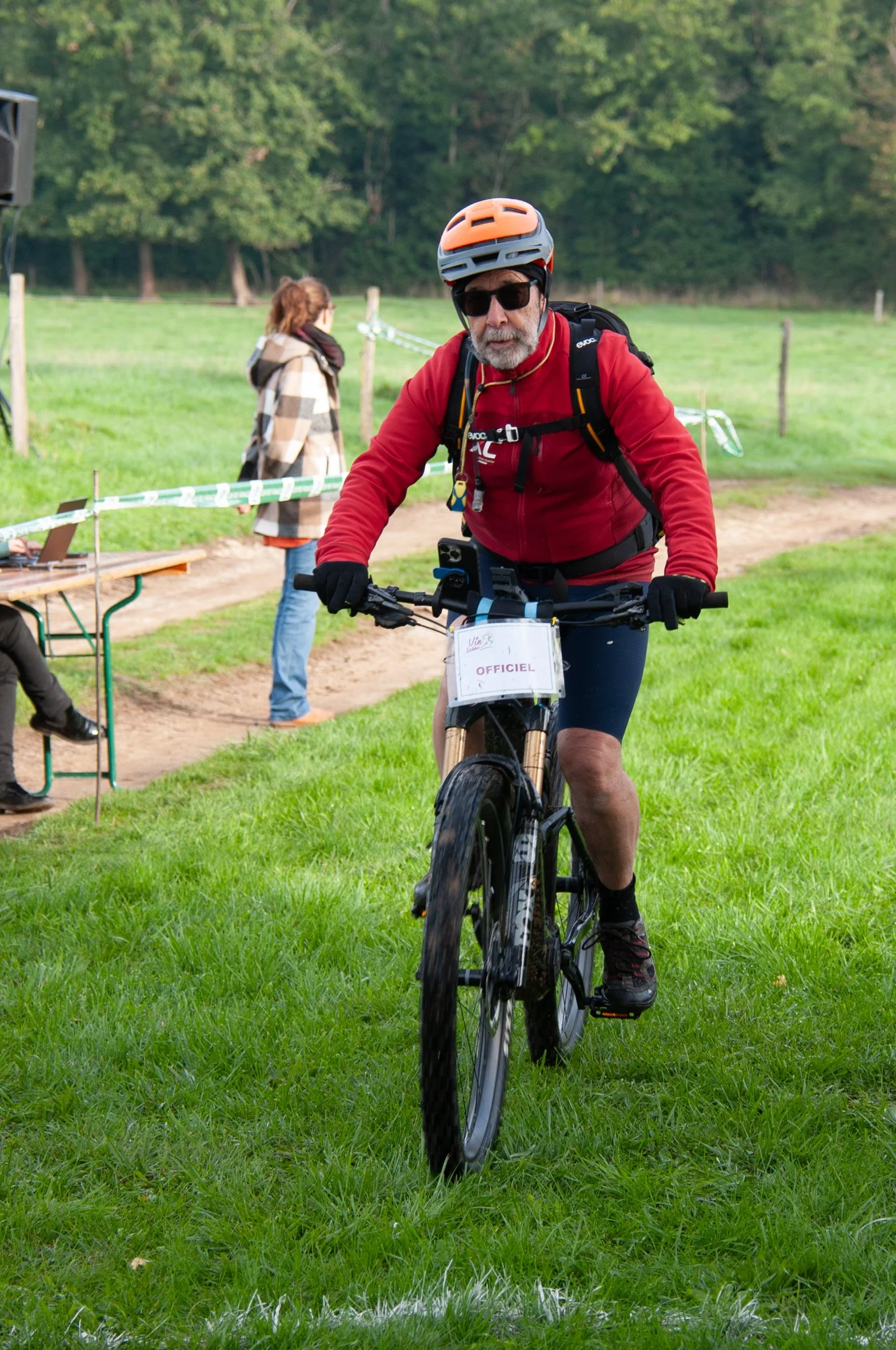 Un homme à vélo électrique portant un casque orange et des lunettes de soleil traverse une zone herbeuse lors d'un événement, avec un panneau indiquant 'OFFICIEL' accroché à la fourche de son vélo, et d'autres personnes en arrière-plan.