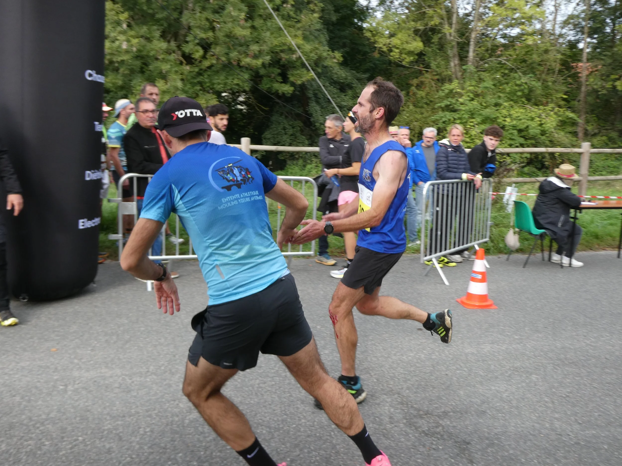 Deux coureurs en pleine course, un homme portant un maillot bleu et un autre en ti-shirt noir et short noir, avec un public derrière les barrières de course et une femme assise à une table au fond.