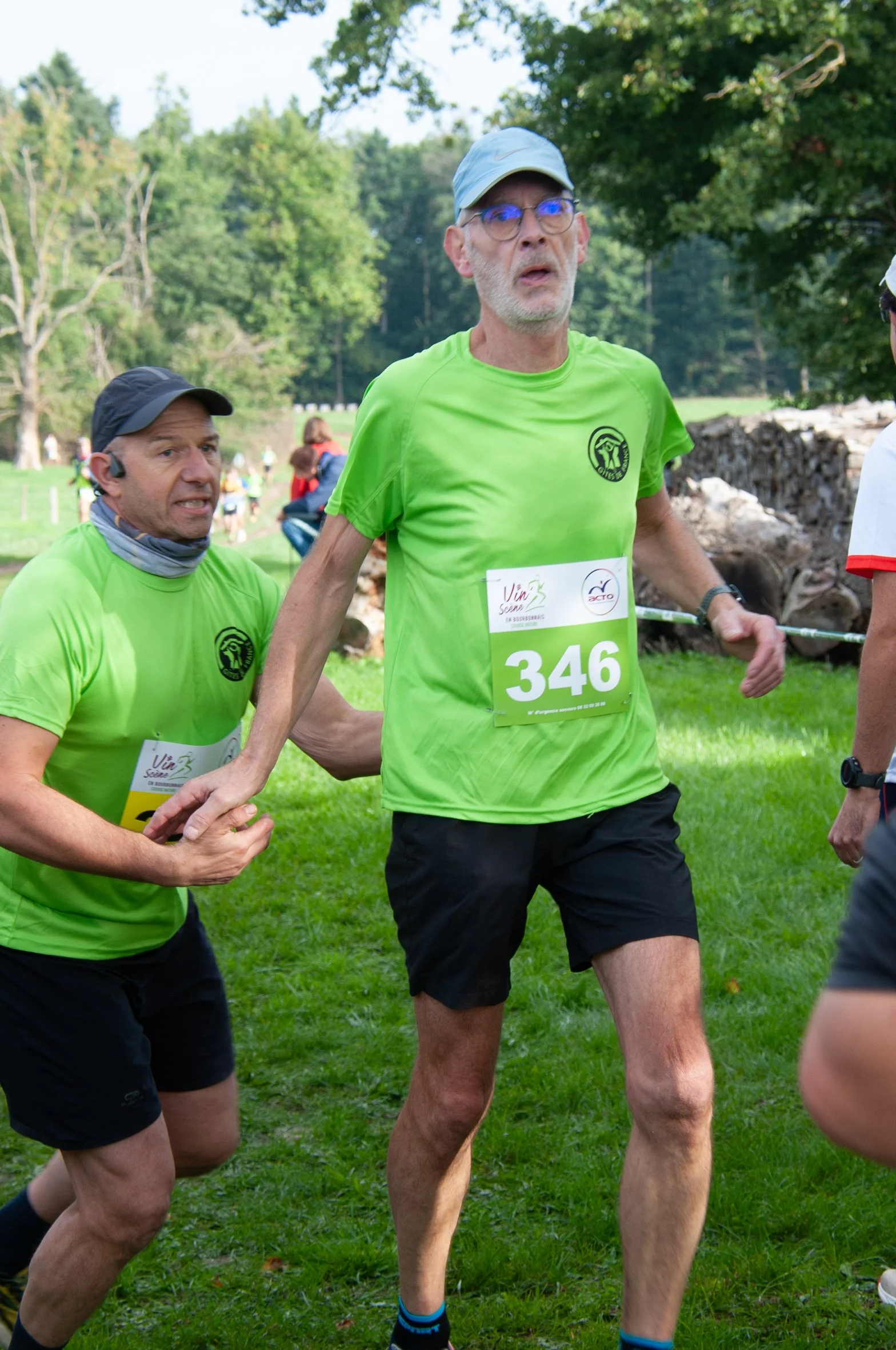 Deux coureurs en tee-shirts verts participant à une course dans un parc verdoyant, avec autres participants en arrière-plan.