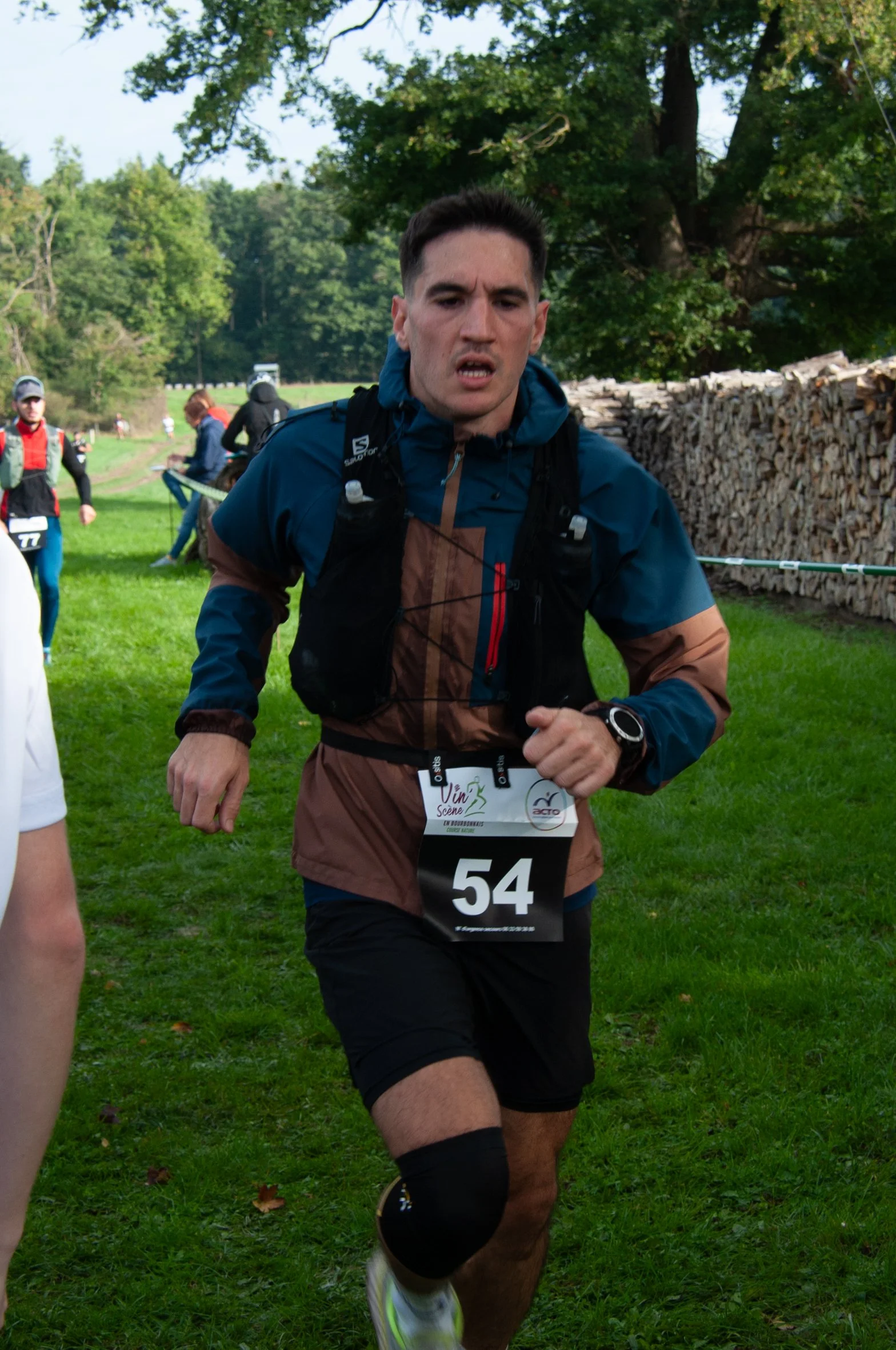 Un homme participe à une course en plein air, portant un dossard numéro 54, vêtu de vêtements de course, avec un genou protégé. Il court dans un espace vert avec des plantes et des arbres en arrière-plan.