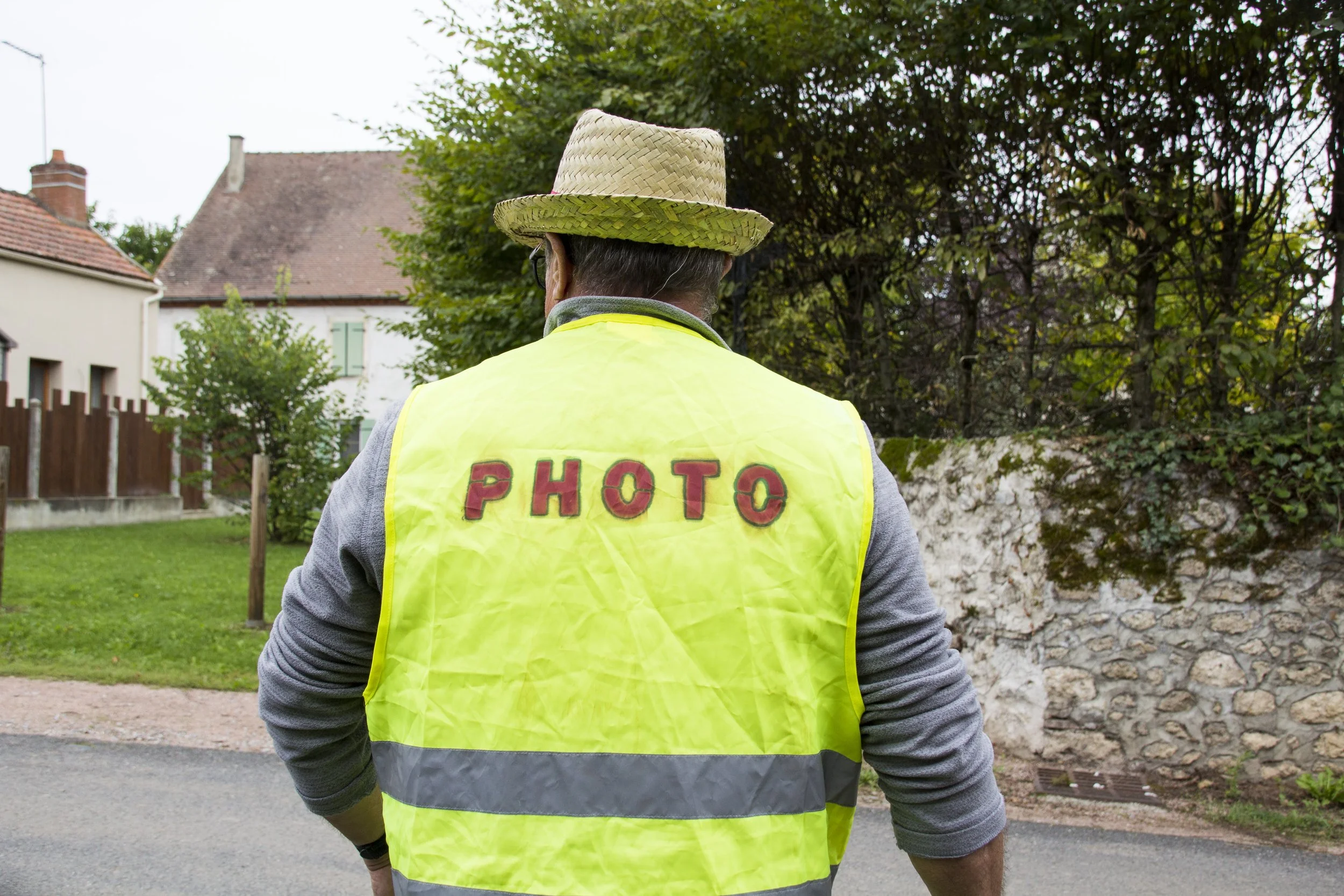 Une personne vue de dos portant une veste jaune avec le mot 'PHOTO' écrit dessus, et un chapeau en paille.
