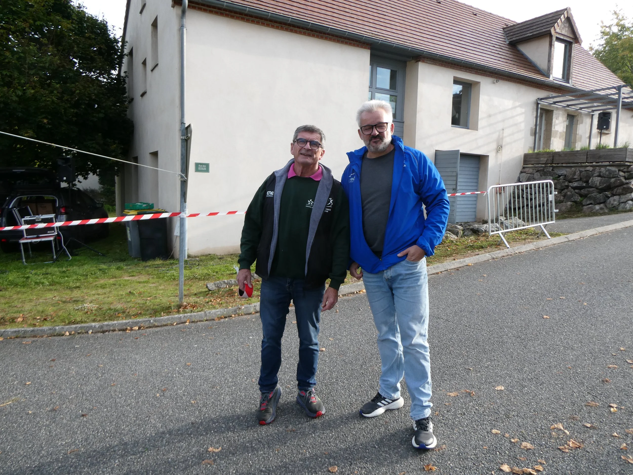 Deux hommes posant ensemble devant une maison, l'un porte un sac à dos et l'autre une veste bleue, dans une rue résidentielle.
