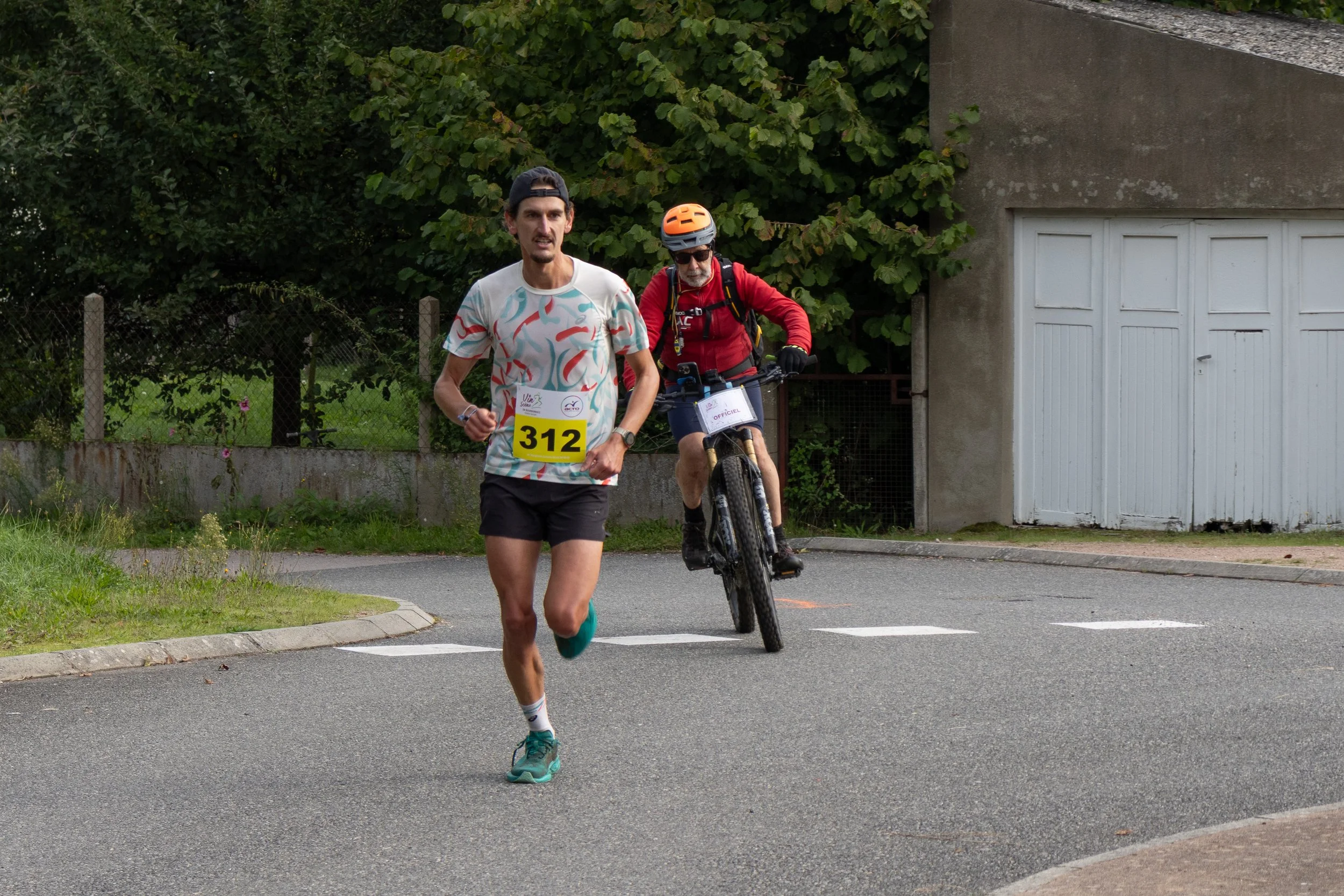 Un homme court sur la route lors d'une course, en arrière-plan un cycliste avec casque et sac à dos, portant le dossard 312. La scène se déroule dans un environnement résidentiel avec des maisons et des arbres.