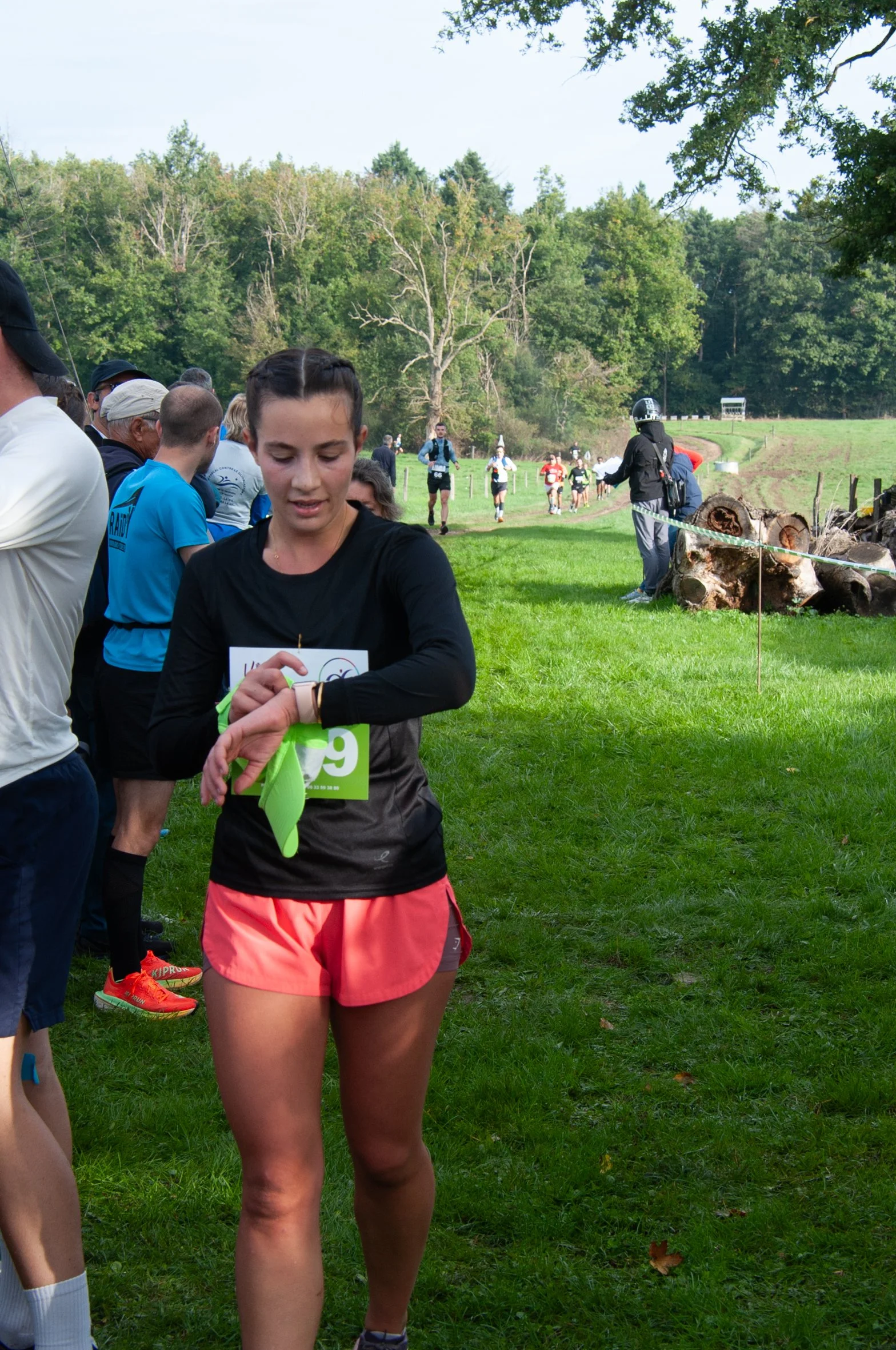 Une jeune femme regarde sa montre lors d'une course dans un parc vert, entourée d'autres coureurs et spectateurs.