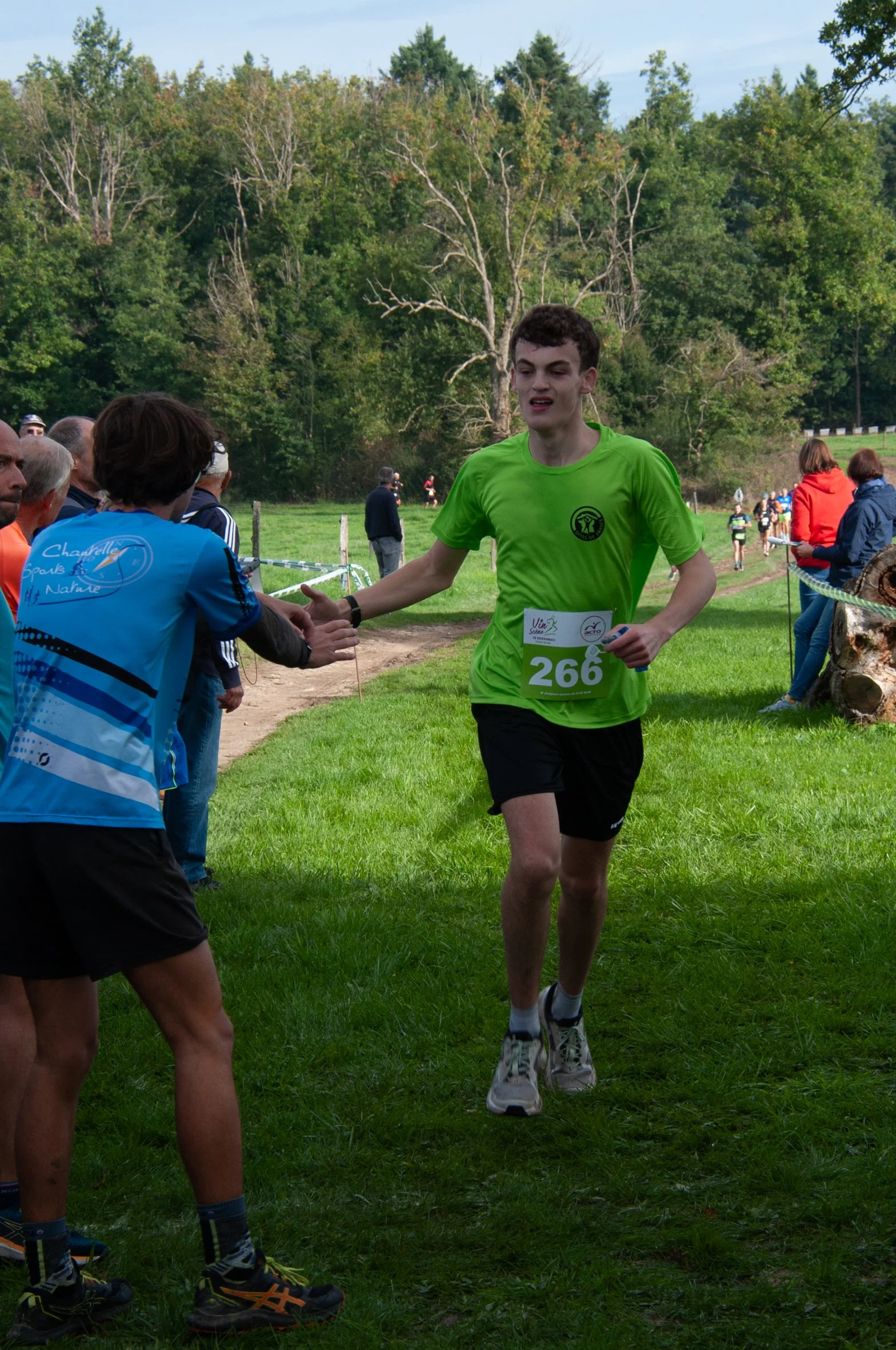Un jeune coureur donne la main à une spectatrice lors d'une course en plein air, avec d'autres coureurs et spectateurs en arrière-plan, sous un ciel partiellement nuageux.