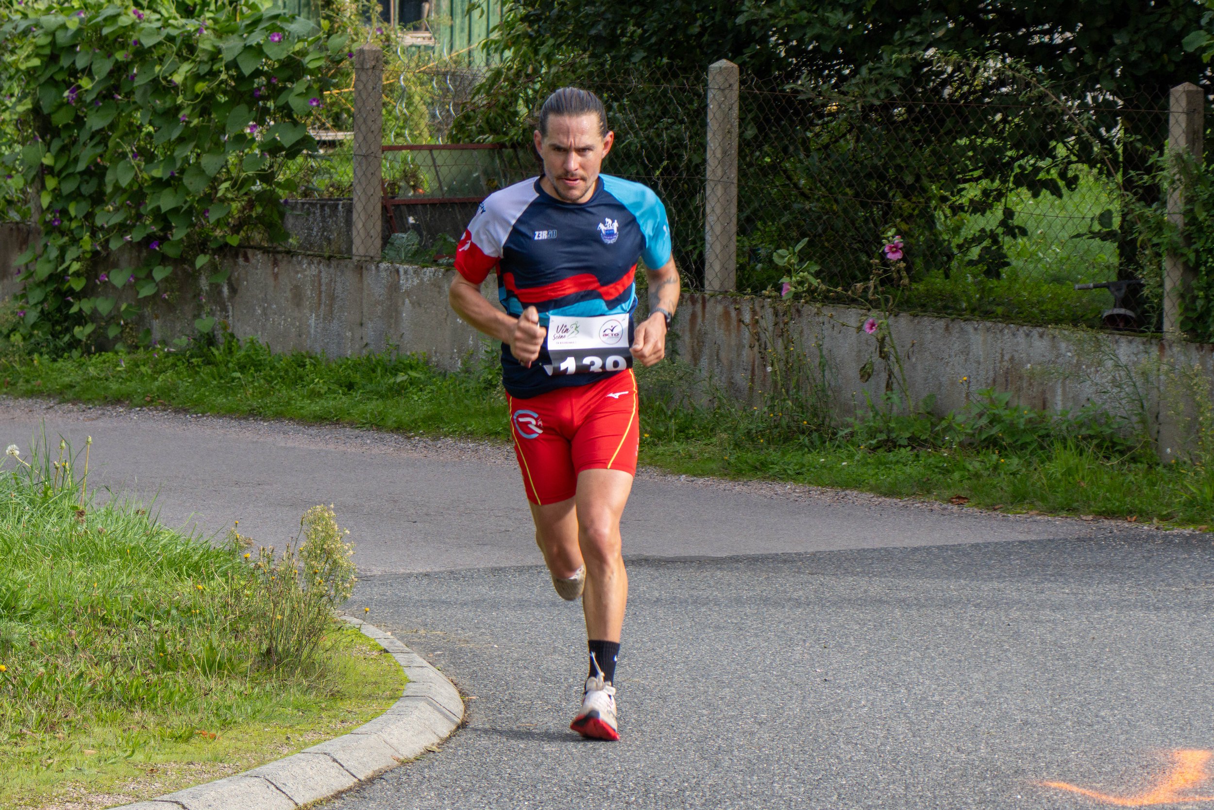 Un homme court sur une route de campagne, il porte un maillot de course bleu, rouge, blanc et une dossard numéro 130.