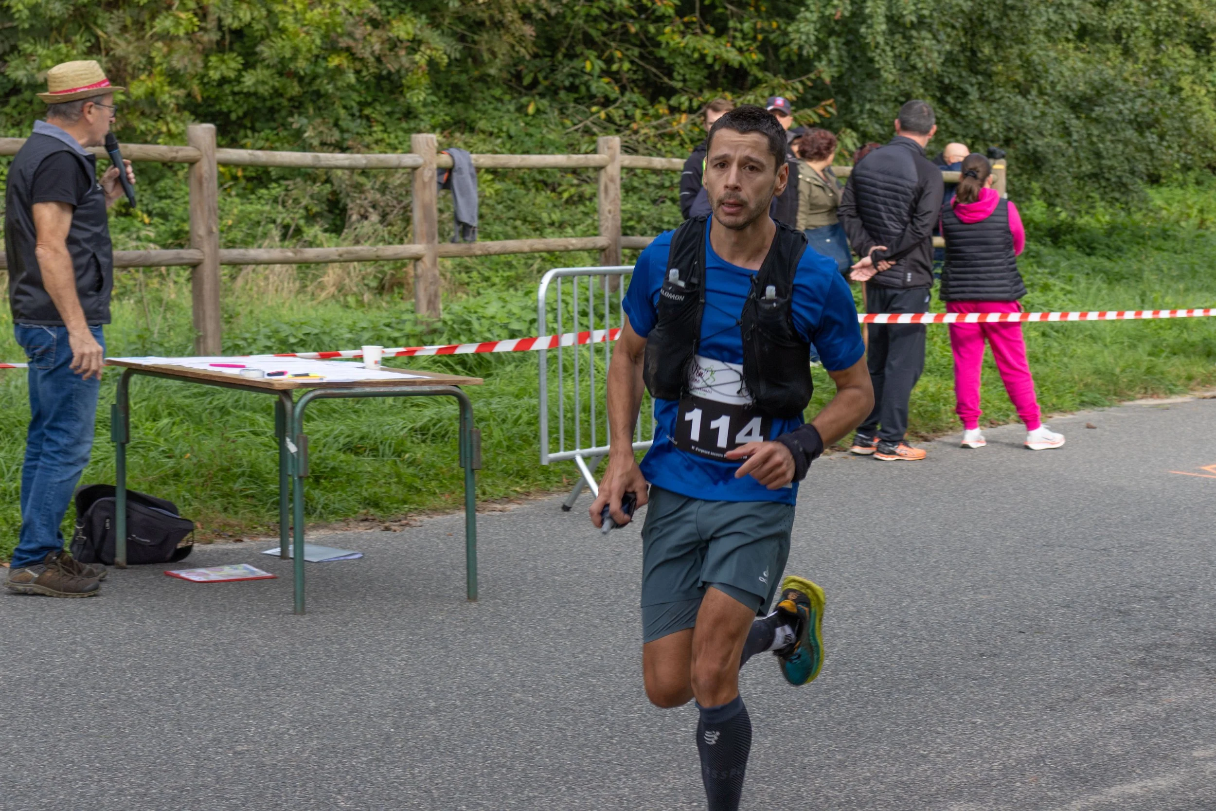 Un homme court lors d'une course, portant un dossard avec le numéro 114, dans un environnement extérieur vert. Il porte une tenue de course bleue, des chaussettes longues noires et des chaussures de sport. Des spectateurs et un officiel sont visibles