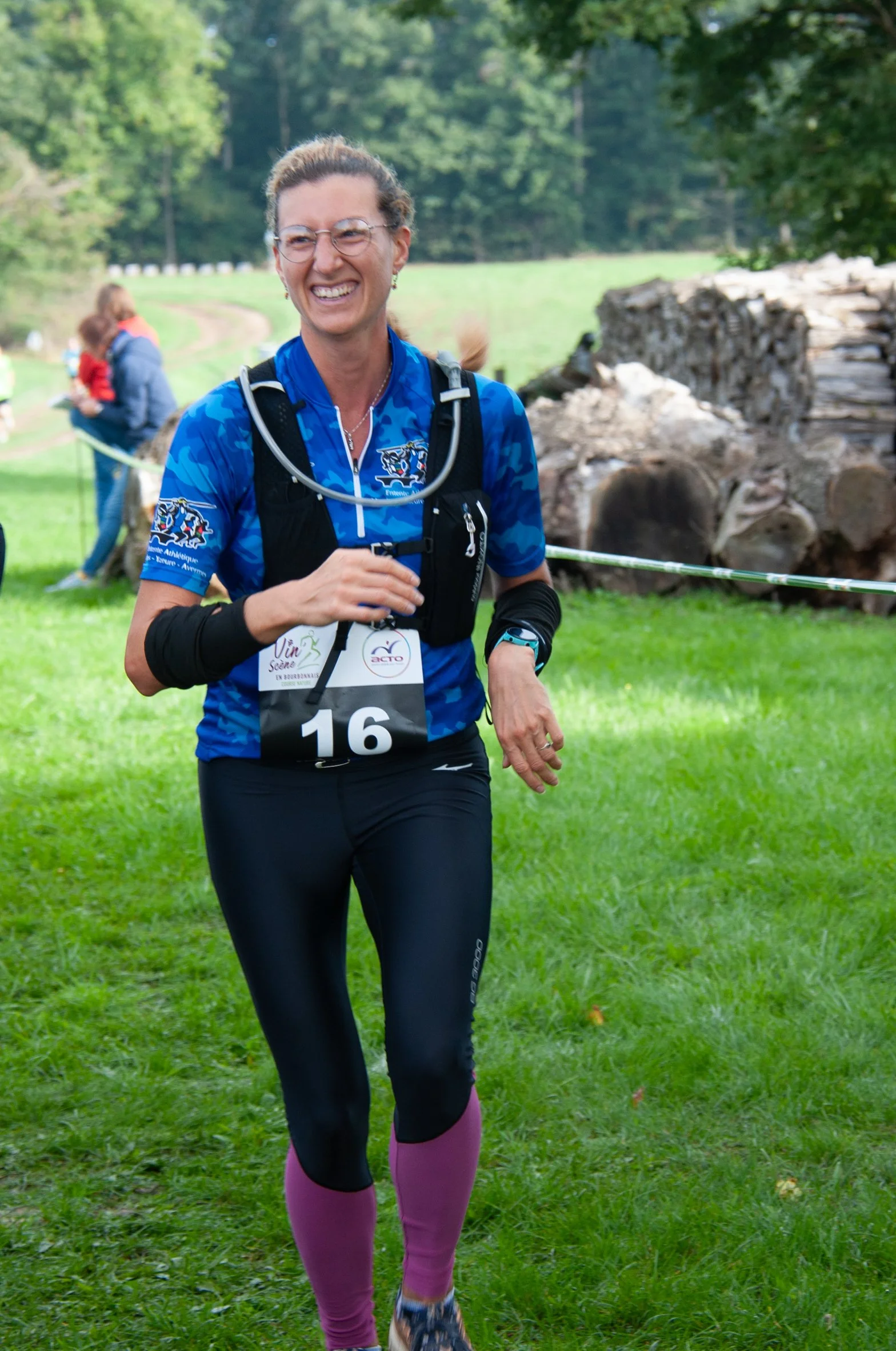 Femme souriante en tenue de course à pied, portant un numéro 16, courant dans un parc verdoyant.