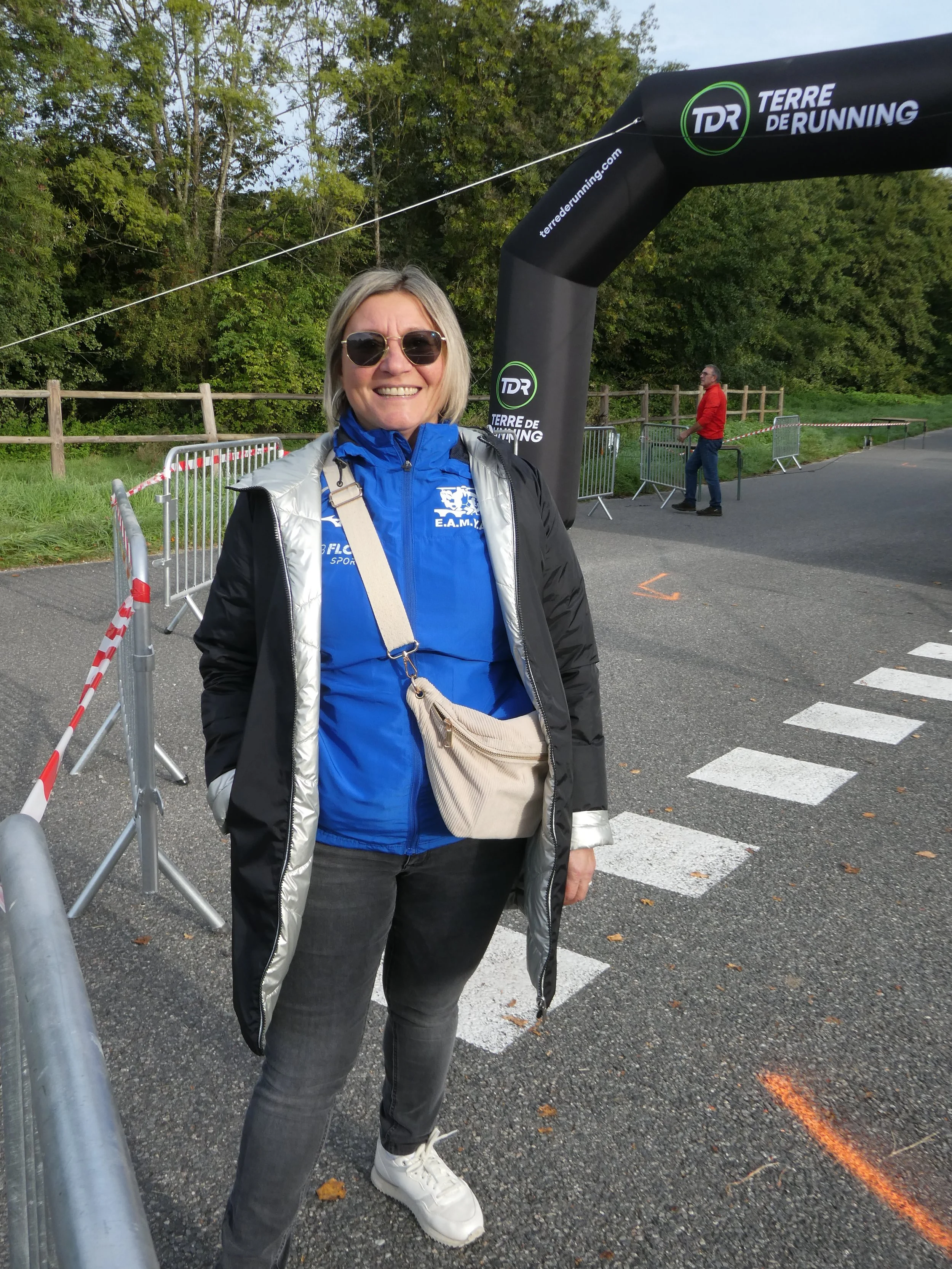 Une femme souriante portant des lunettes de soleil, un blouson noir et gris, une veste bleue, un sac beige, et des chaussures blanches, debout à côté d'une barrière métallique à une course ou un événement sportif, avec un grand arc gonflable noir mar