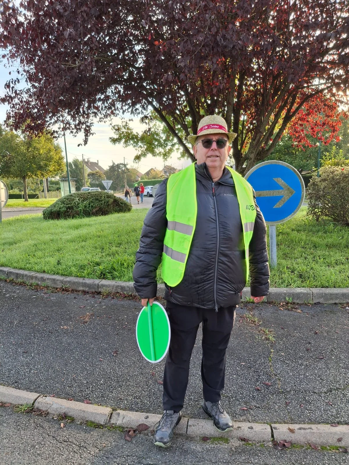 Homme en veste noire et gilet jaune vif avec un chapeau, des lunettes de soleil, debout sur un trottoir avec un panneau de signalisation routière en arrière-plan.
