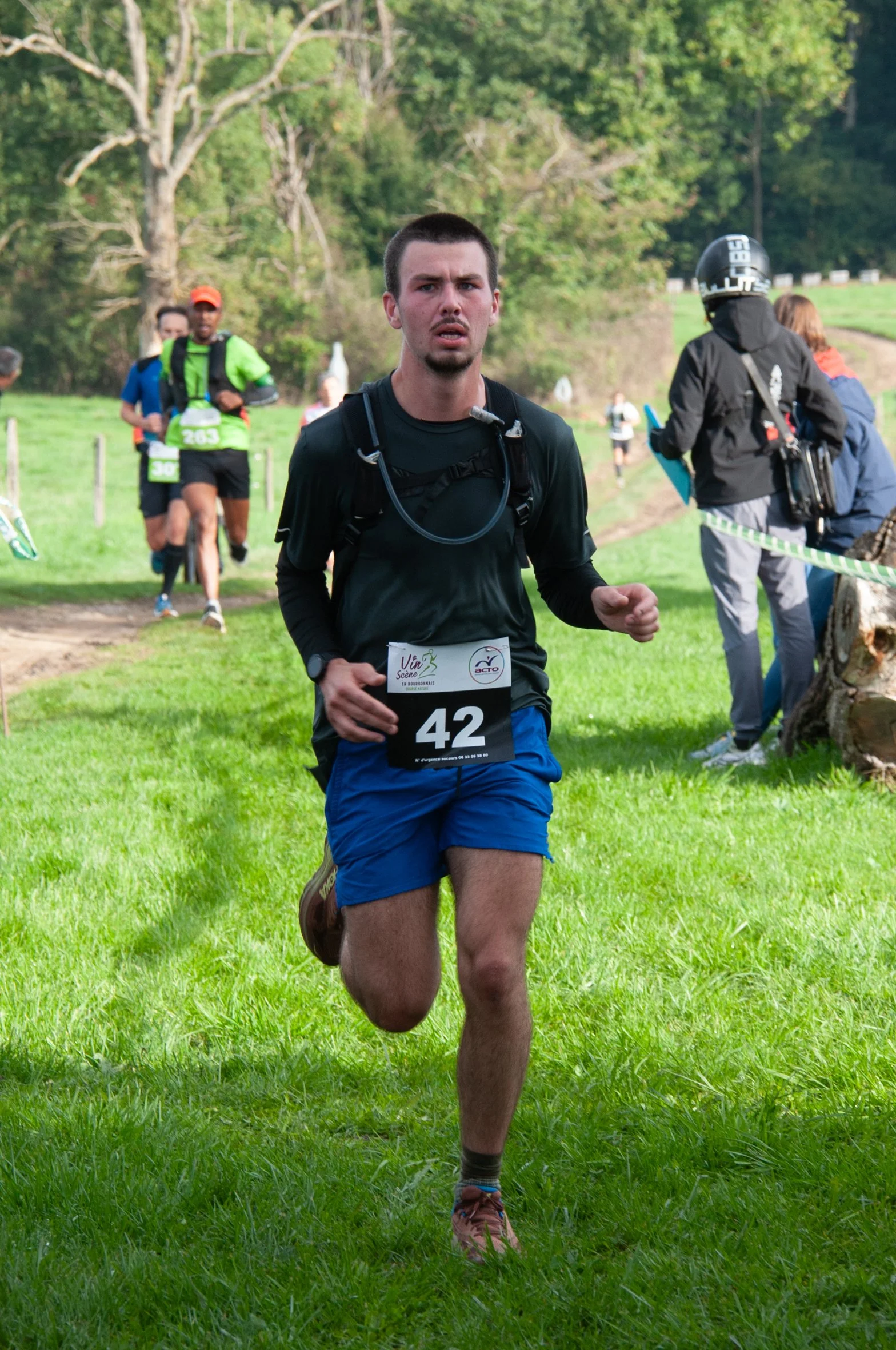 Un homme court dans une course en plein air, portant un dossard avec le numéro 42, vêtu d'un t-shirt noir, un short bleu et des chaussures de course. Il y a d'autres coureurs et spectateurs en arrière-plan, dans un environnement verdoyant avec des ar