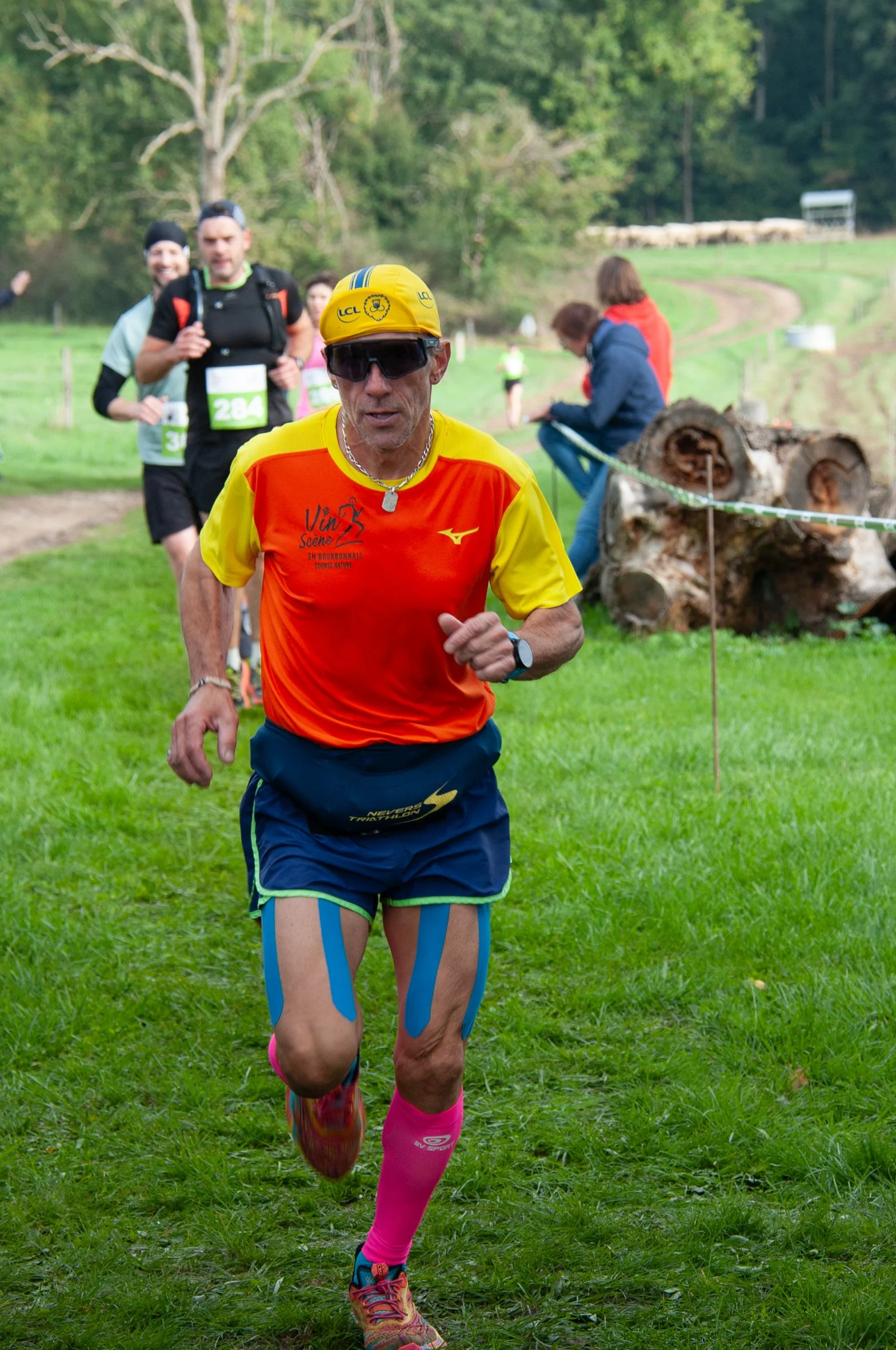 Un homme en course à pied portant un casque jaune, des lunettes de soleil, un maillot orange et jaune, des shorts bleus, des chaussettes roses, et des chaussures de course, lors d'une compétition dans un environnement herbeux avec d'autres coureurs e