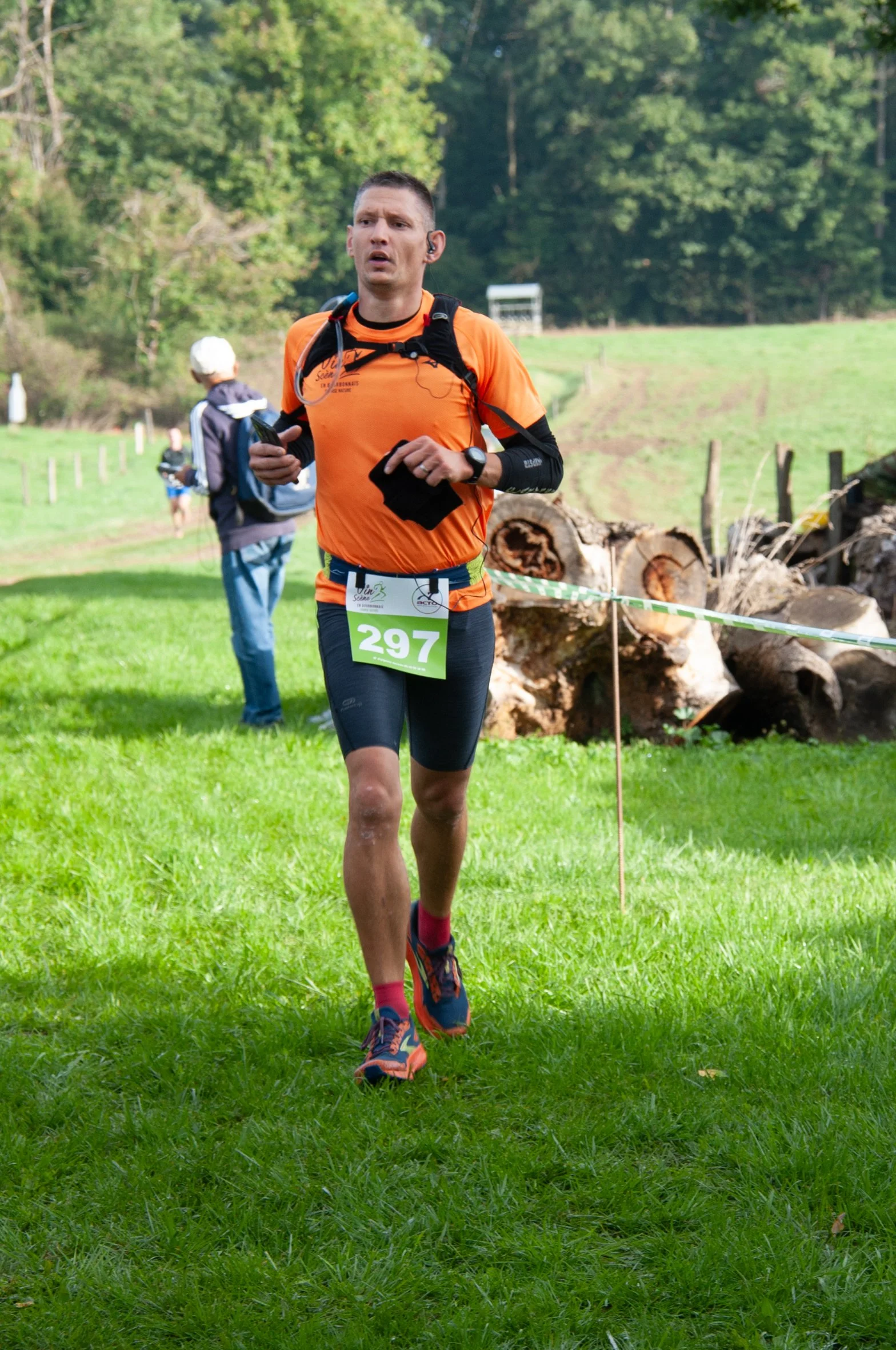 Un homme court lors d'une course en plein air, portant un t-shirt orange, un maillot de course noir, un numéro 297, une montre de sport, et des écouteurs, dans un environnement verdoyant avec des arbres et des troncs de bois en arrière-plan.