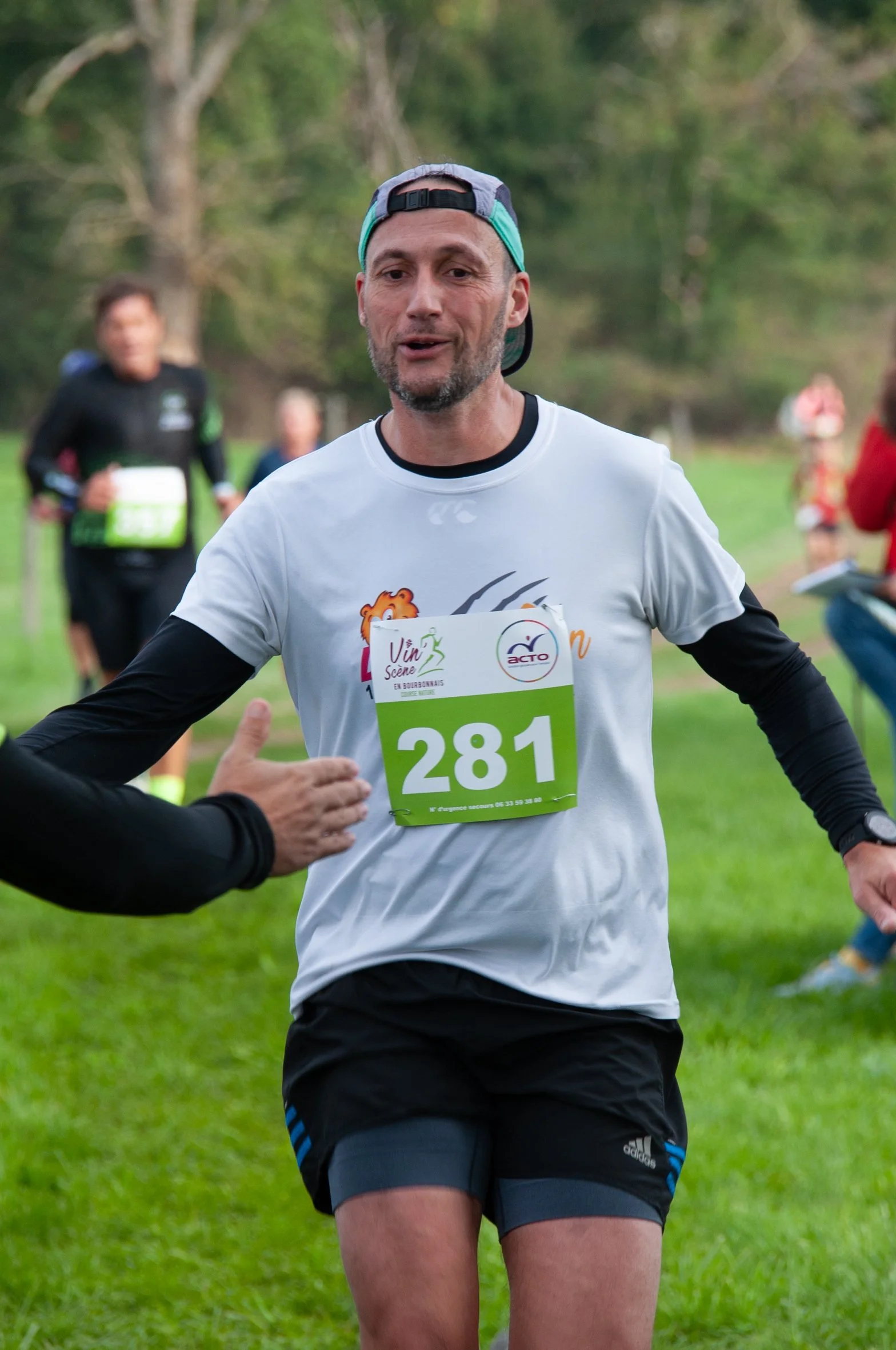 Un homme participe à une course en plein air, portant un numéro de dossard 281, vêtu d'un t-shirt blanc, d'un bonnet déóarlé et de shorts noirs, serrant la main d'une autre personne, avec d'autres coureurs en arrière-plan.