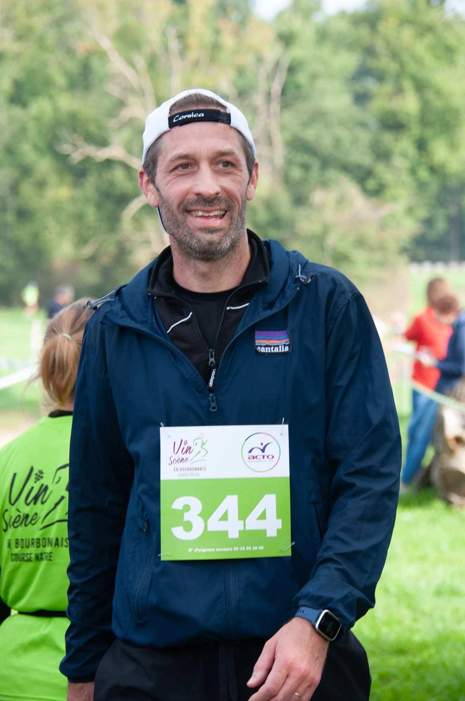 Un homme souriant portant un dossard numéro 344 lors d'une course en plein air, avec des personnes en arrière-plan dans un parc verdoyant.