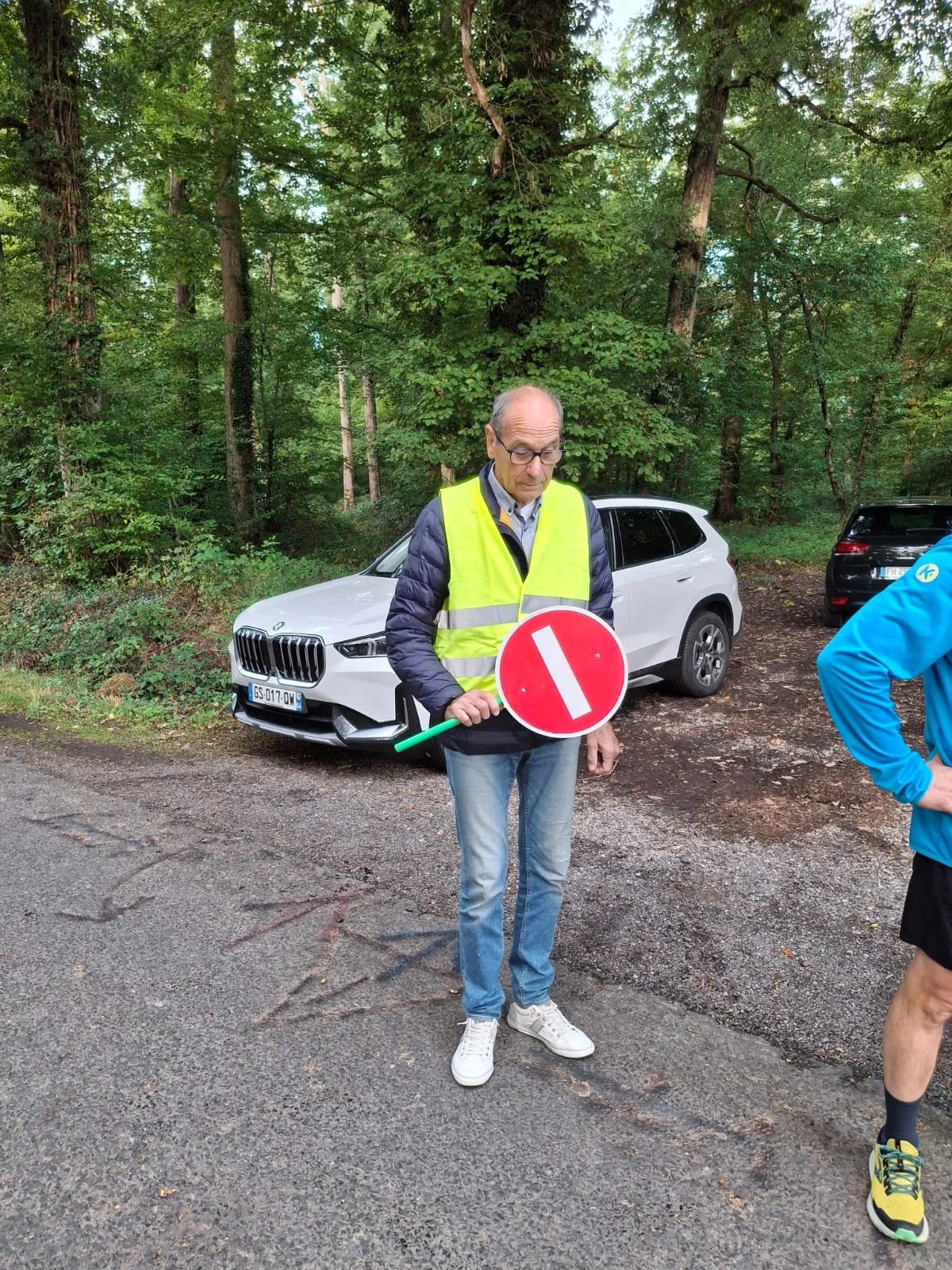 Un homme portant un gilet jaune réfléchissant tient un panneau d'interdiction à la main, se tenant sur une route en forêt avec plusieurs voitures en arrière-plan.