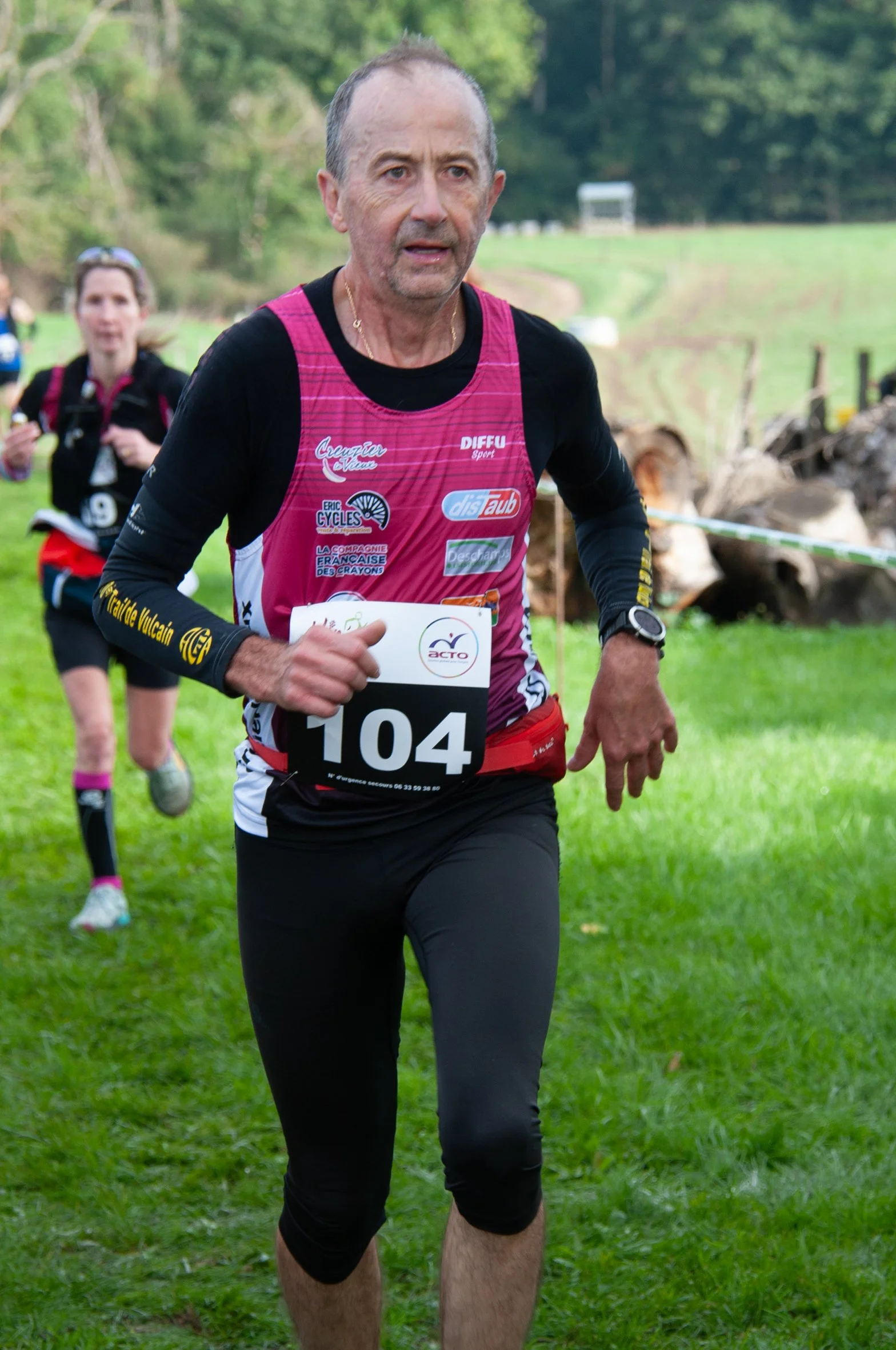 Un homme court lors d'une course en plein air, portant un t-shirt noir avec un maillot de course rose. Il a un numéro 104 attaché à sa tenue. En arrière-plan, d'autres participants courent dans un environnement verdoyant.