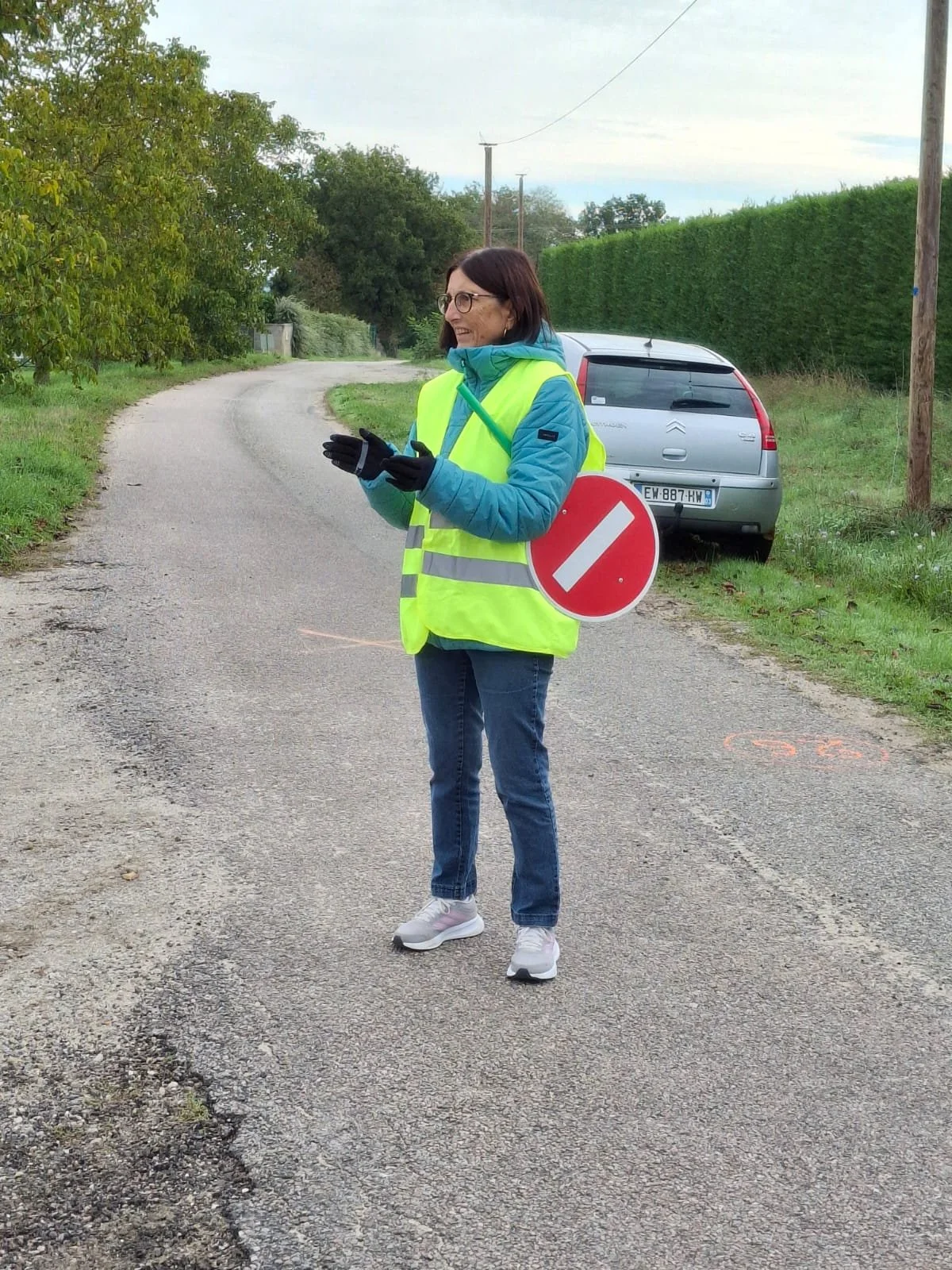 Une femme portant un gilet de sécurité jaune et un gilet bleu, se tenant au milieu d'une route rurale, tenant un panneau de signalisation interdit. Elle porte également des gants noirs, des lunettes et des chaussures blanches, avec une voiture grise 