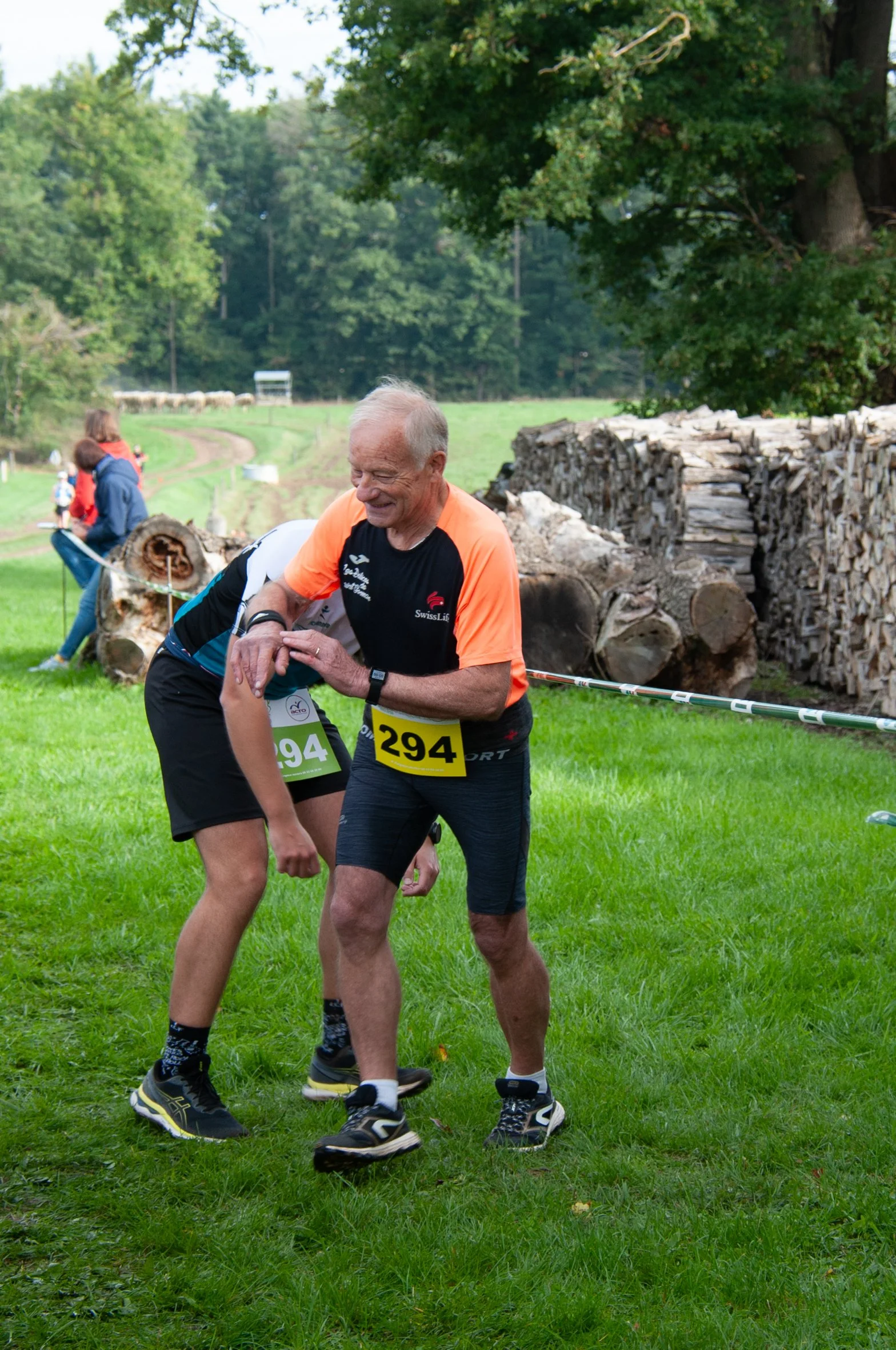 Un homme âgé en tenue de course, portant le numéro 294, sourit et semble aider ou encourager un jeune coureur lors d'une course en plein air dans une zone verdoyante avec des arbres et du bois empilé en arrière-plan.