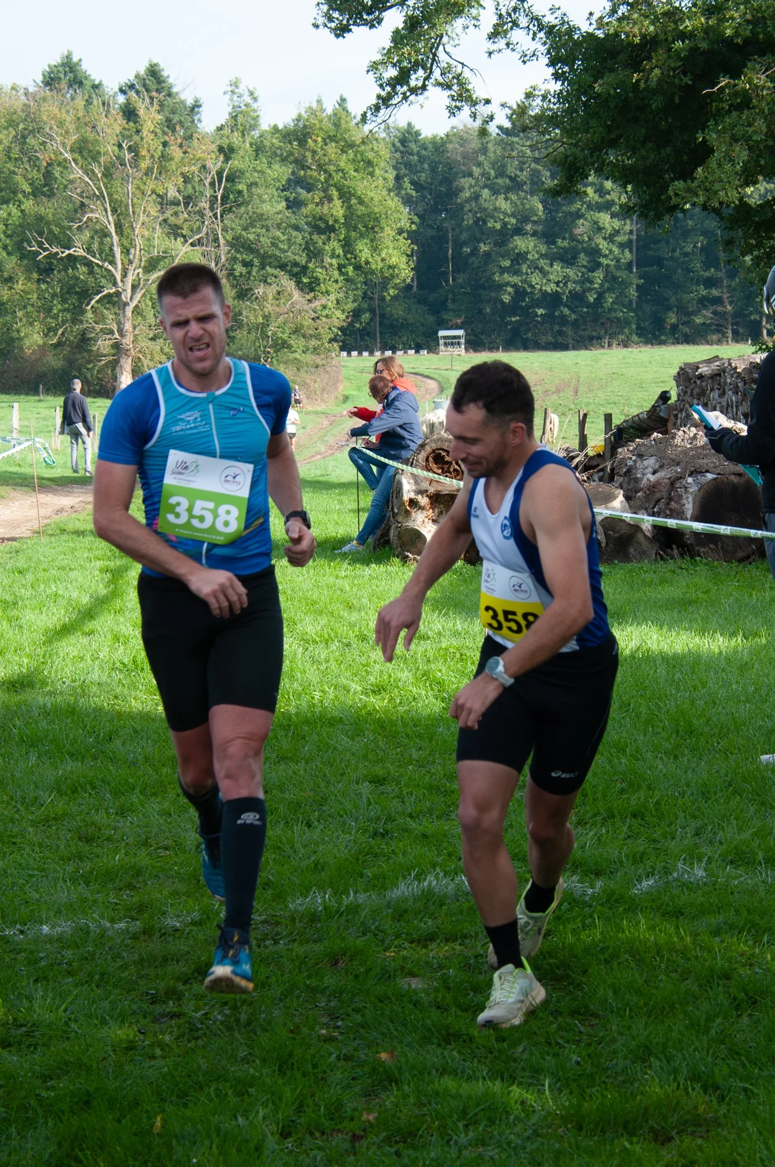 Deux coureurs participent à une course en plein air, portant des dossards numérotés 358 et 359. L'un porte un t-shirt bleu et l'autre un débardeur blanc et bleu, tous deux en pleine marche sur l'herbe verte.