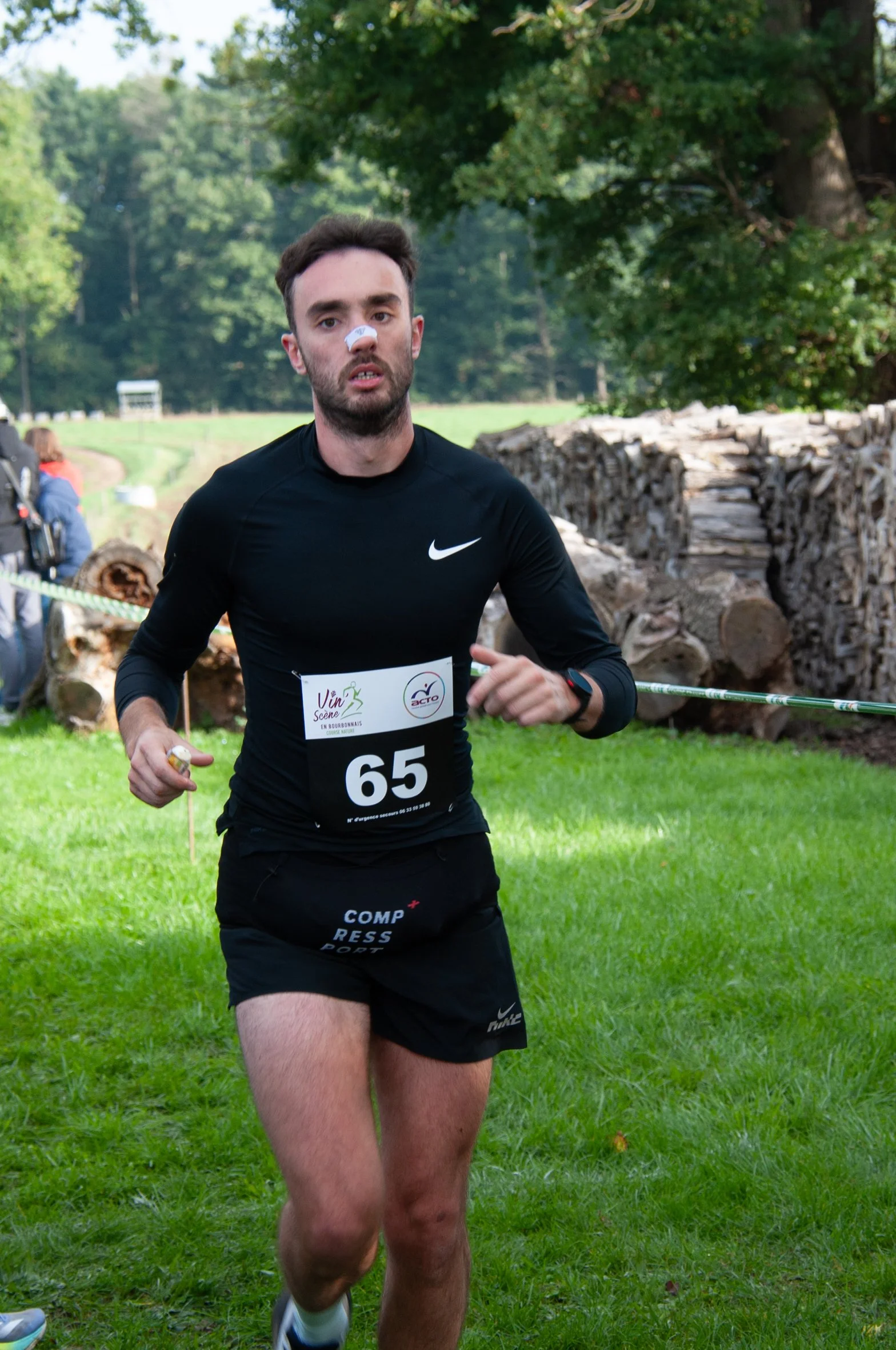 Homme courant lors d'une compétition, portant un maillot noir avec le logo Nike, un numéro 65, et une bande adhésive blanche sur le nez, dans un environnement verdoyant avec des arbres et des piles de bois.