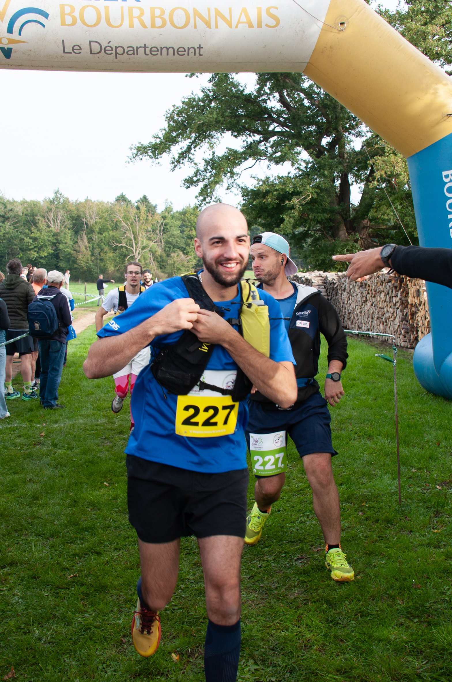 Deux hommes courent lors d'une course en plein air, portant des dossards numérotés 227. Le premier homme est souriant et porte un t-shirt bleu, le deuxième porte une casquette et un t-shirt noir avec un short. Il y a d'autres participants en arrière-