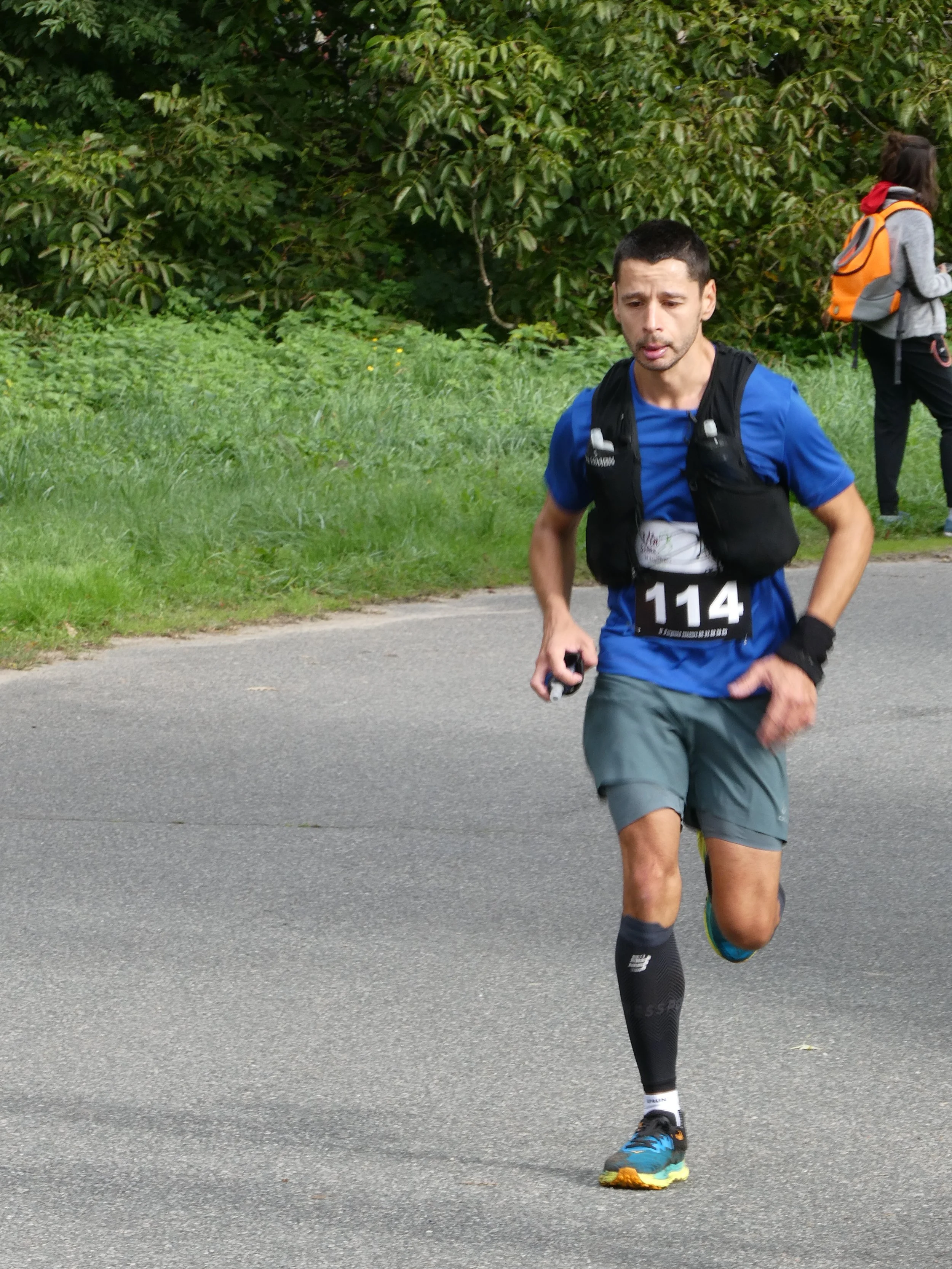 Homme courant lors d'une course en extérieur, portant un numéro de dossard 114, un t-shirt bleu, un short gris, une chaussure de course bleu et noir, et un brassard noir, avec un pack à eau sur le dos, en train de courir sur une route avec des arbres
